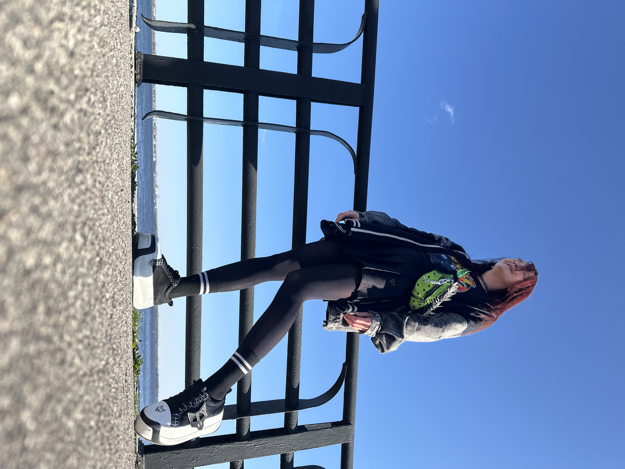 The image, taken in Saratov, Russia, features a young person posing outdoors on a bright, sunny day. The individual, appearing to be a young woman with reddish-brown hair, is captured leaning against a black metal railing. She wears a dark denim jacket over a black top or dress, black leggings, and black socks with white stripes. Her footwear consists of distinctive chunky black and white platform sneakers, with the brand "NAKED WOLFE" visible on the side of one shoe. A vibrant green and yellow patterned bag or scarf is draped over her chest.
The scene is set on what appears to be a paved promenade or walkway, with a coarse, granular texture underfoot. Beyond the railing, a calm body of water stretches into the distance, with a faint shoreline or landmass visible on the horizon. The sky above is a clear, brilliant blue, showing only a single small white cloud. The person's head is tilted slightly upwards, suggesting she is looking towards the sky or enjoying the bright weather. The black metal railing features a grid-like design with curved support elements. The overall impression is one of relaxed outdoor activity under pleasant weather conditions.