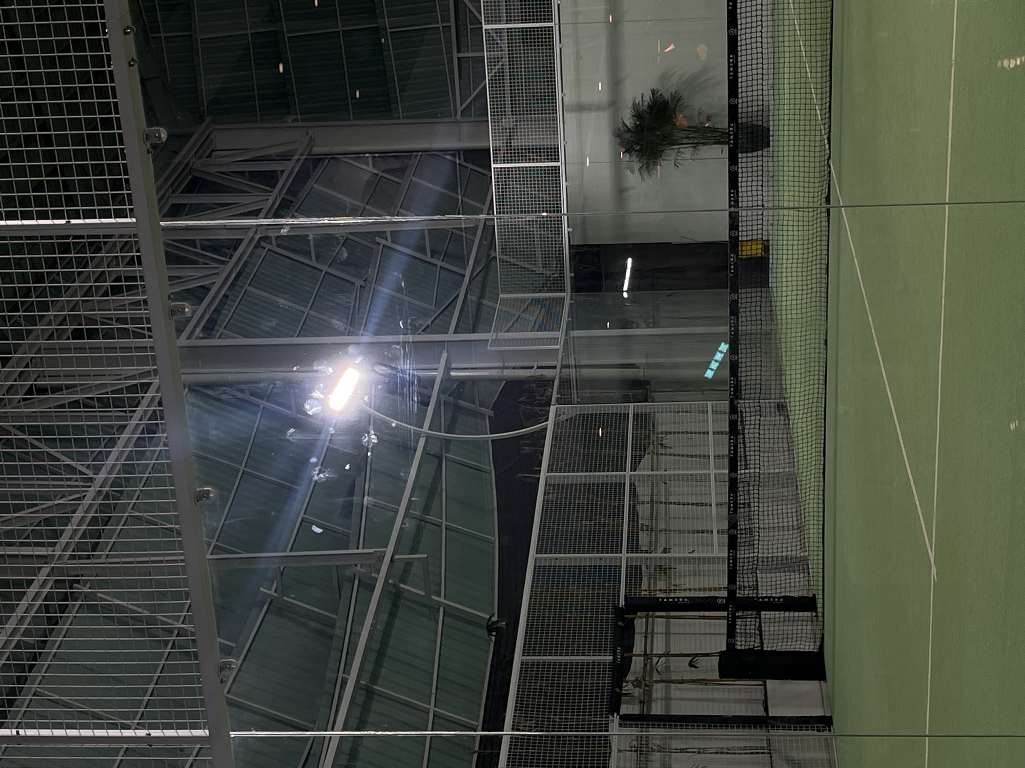 The image depicts an indoor padel tennis court. The court is green with white lines. A net divides the court. Metal mesh fencing surrounds the court. The building has a high, industrial-style ceiling with exposed metal beams and a translucent roof panel. A spotlight illuminates the space. A potted plant is visible along the side of the court. The image was taken in North Kuta, Indonesia.