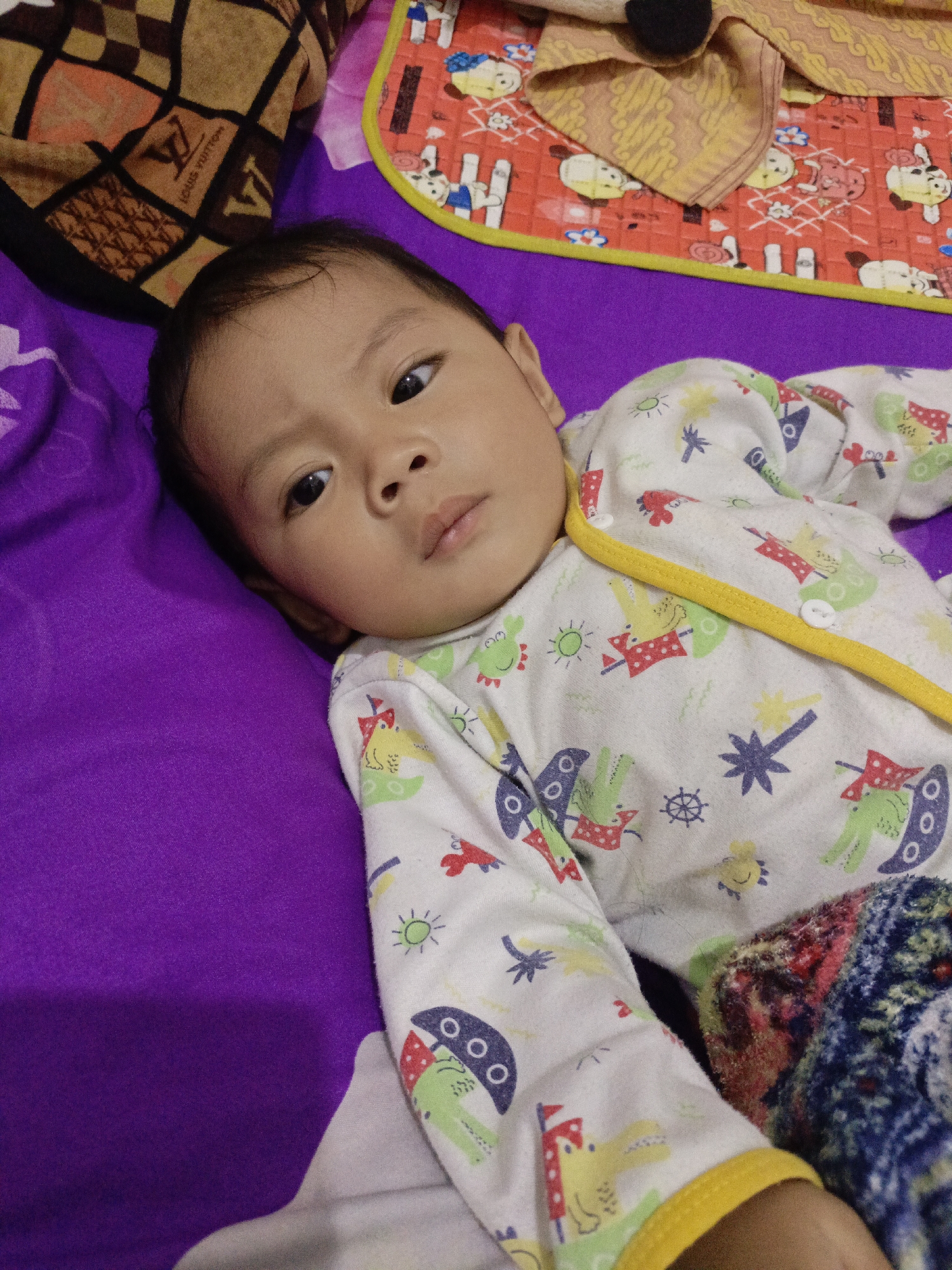 A young baby lies on a purple surface, likely a bed or playmat. The baby is wearing a long-sleeved onesie with a playful pattern of cartoon animals, including alligators, fish, and suns, in various colors like green, yellow, red, and blue. The onesie has a yellow trim around the neckline and cuffs, and a white button at the collar. The baby's facial expression is neutral, with their eyes open and looking towards the camera.
In the background, to the left of the baby, a brown blanket with a repeating pattern of the Louis Vuitton logo is visible. To the right of the baby, a red and yellow patterned mat or blanket is partially visible. The overall lighting suggests it is daytime, possibly indoors. The scene is confined to the baby and the immediate surroundings, with no clear indications of weather or specific location beyond the general context of a domestic setting in Pager, Indonesia. The focus is entirely on the baby.