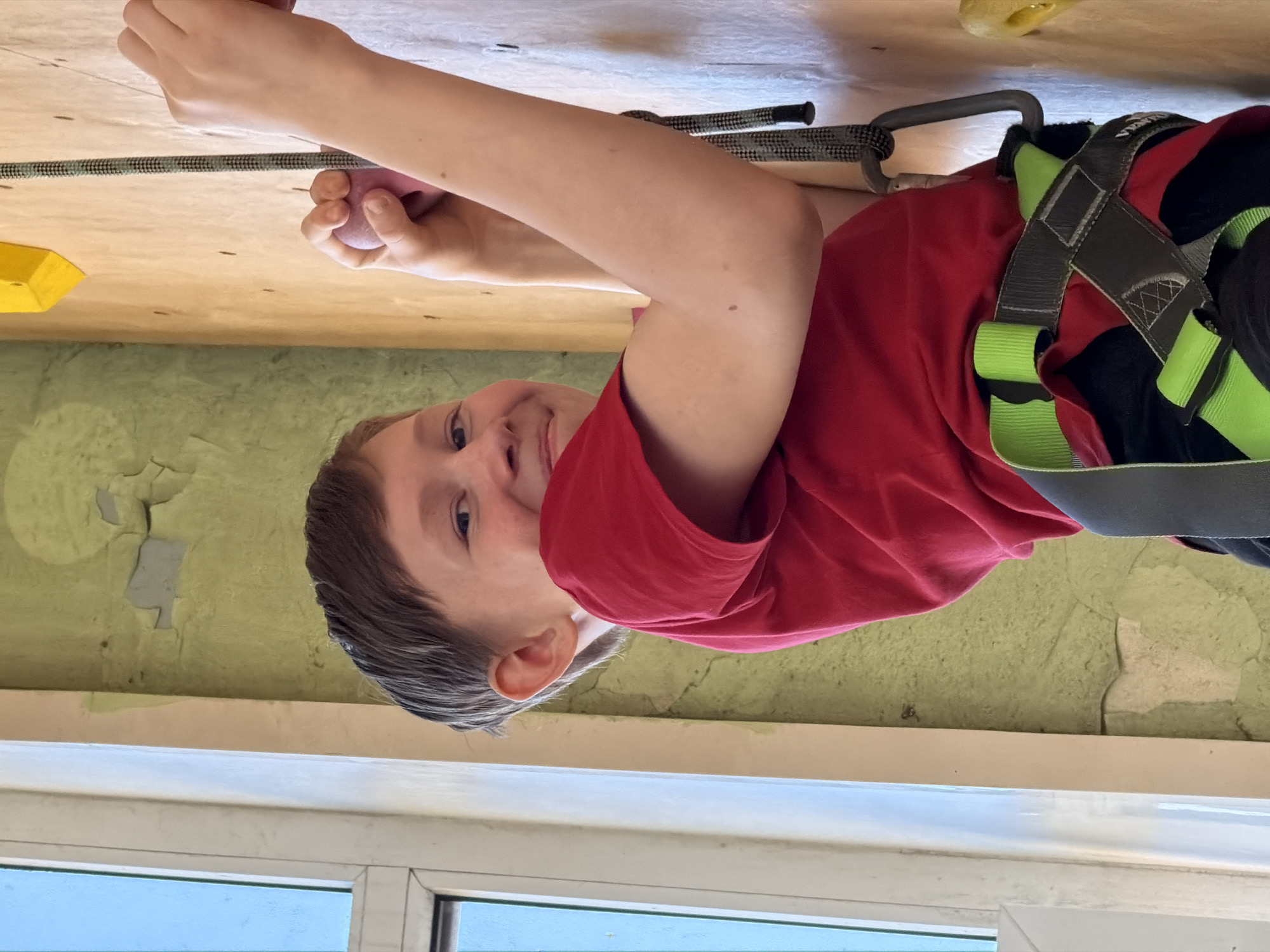 A young boy is captured in an active pose, seemingly climbing or hanging on an indoor climbing wall. He is smiling cheerfully and looking directly at the camera, conveying a sense of enjoyment. He wears a bright red t-shirt and a climbing harness, which is dark grey with striking lime green accents. A patterned grey and black climbing rope is attached to his harness via a silver carabiner, indicating safety precautions are in place. His right hand firmly grips a pink, rounded climbing hold, while other holds, including a yellow block-shaped one, are visible on the light-colored wall surface.
The background features a light green wall with noticeable patches of peeling paint, suggesting an older or well-used facility. At the bottom of the image, a white window frame reveals a clear blue sky or distant view, indicating the photo was taken during daylight hours. The overall scene depicts a moment of playful exertion and happiness in an indoor climbing environment in Izhevsk, Russia. No visible text can be discerned in the image.
