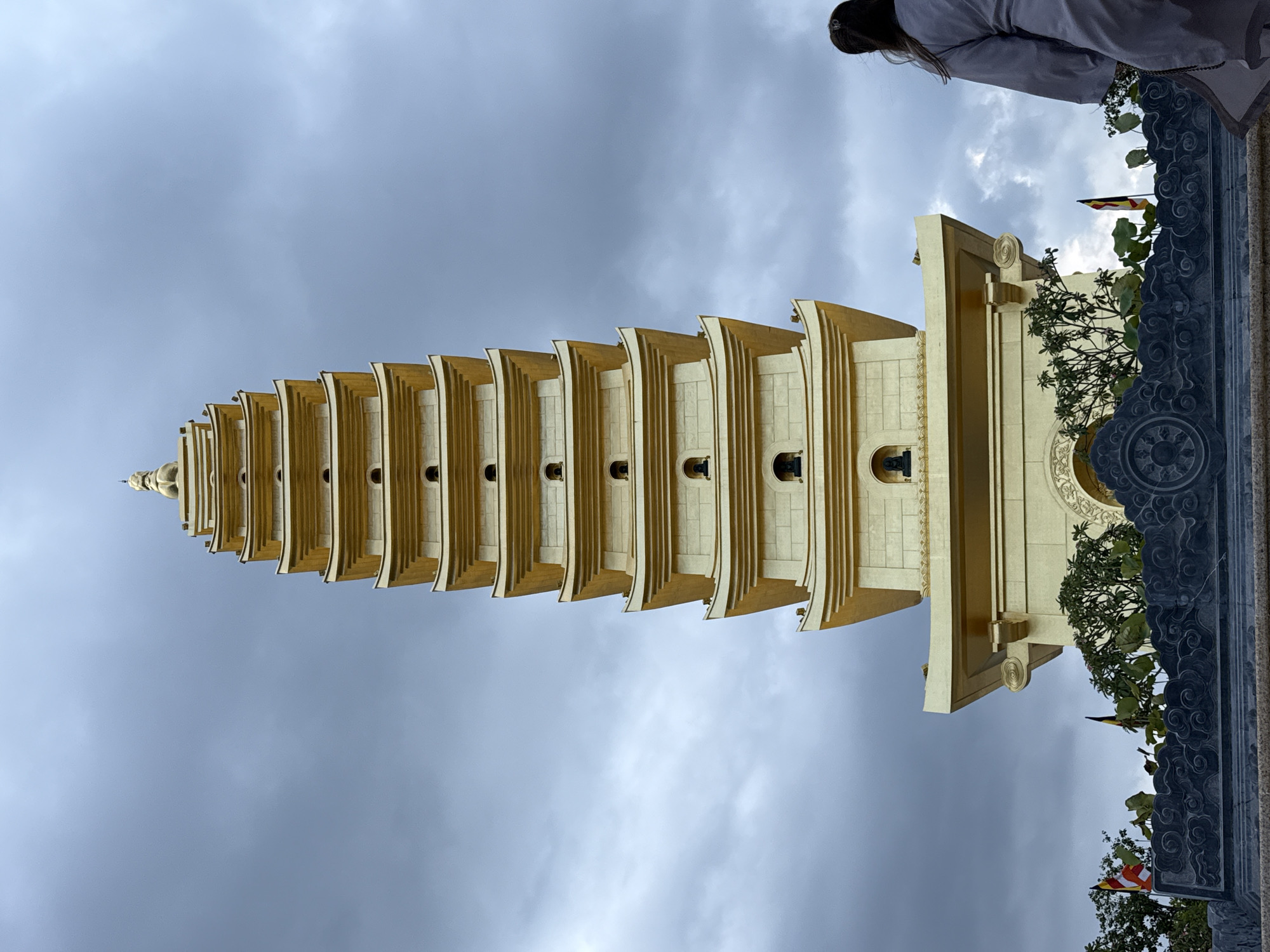 A vertical eye-level shot depicts the gleaming golden pagoda of the Khánh An Monastery in Diên Khánh, Vietnam, towering against an overcast sky. The pagoda's architecture features multiple tiers, diminishing in size as they rise, each with projecting eaves. Ornate details can be seen on the base tier, with carved stonework, foliage, and a small flag visible. The sky is a canvas of gray clouds. A person, seen from the shoulder up, appears in the upper right corner, clad in a white garment, positioned beside an ornately carved ledge.