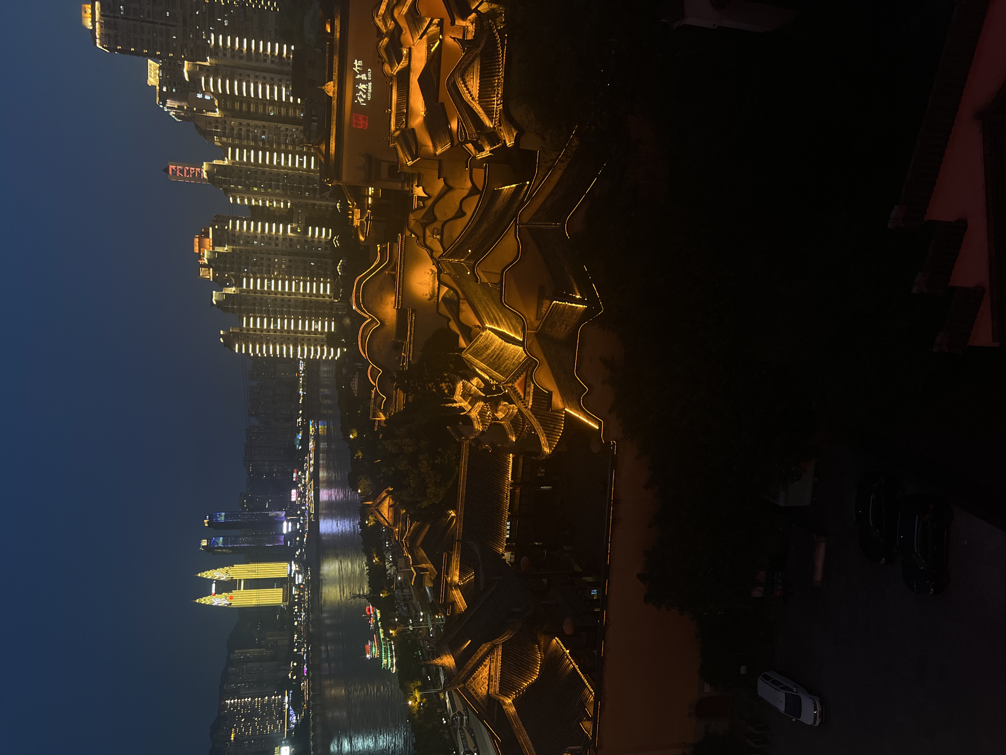 The image captures a night view of the Yuzhong District in Chongqing, China, showcasing a blend of traditional and modern architecture along the waterfront. In the foreground, a cluster of buildings with intricately curved, dark tiled roofs are illuminated by warm, golden lights outlining their edges. These structures appear to be part of a historical district or a themed entertainment area, with ornate details and several layers of roofs visible.
Beyond this traditional architecture, a wide river reflects the lights of the city. On the opposite bank, a dense cityscape of modern high-rise buildings rises, many of which are lit up with rows of bright white lights. Dominating the skyline are several prominent towers, including a pair of striking, golden-lit skyscrapers with pointed tops, resembling twin needles. Other taller buildings display a mix of illuminated windows and architectural lighting.
Further in the distance, the silhouettes of hills or mountains can be seen against the dark night sky. Closer to the foreground, a few vehicles are parked on a street below the traditional buildings, including a white limousine and two dark sedans. The overall scene is one of a vibrant, illuminated city at night, with the contrast between the old and new architectural styles being a prominent feature. The lighting creates a dramatic and somewhat ethereal atmosphere.