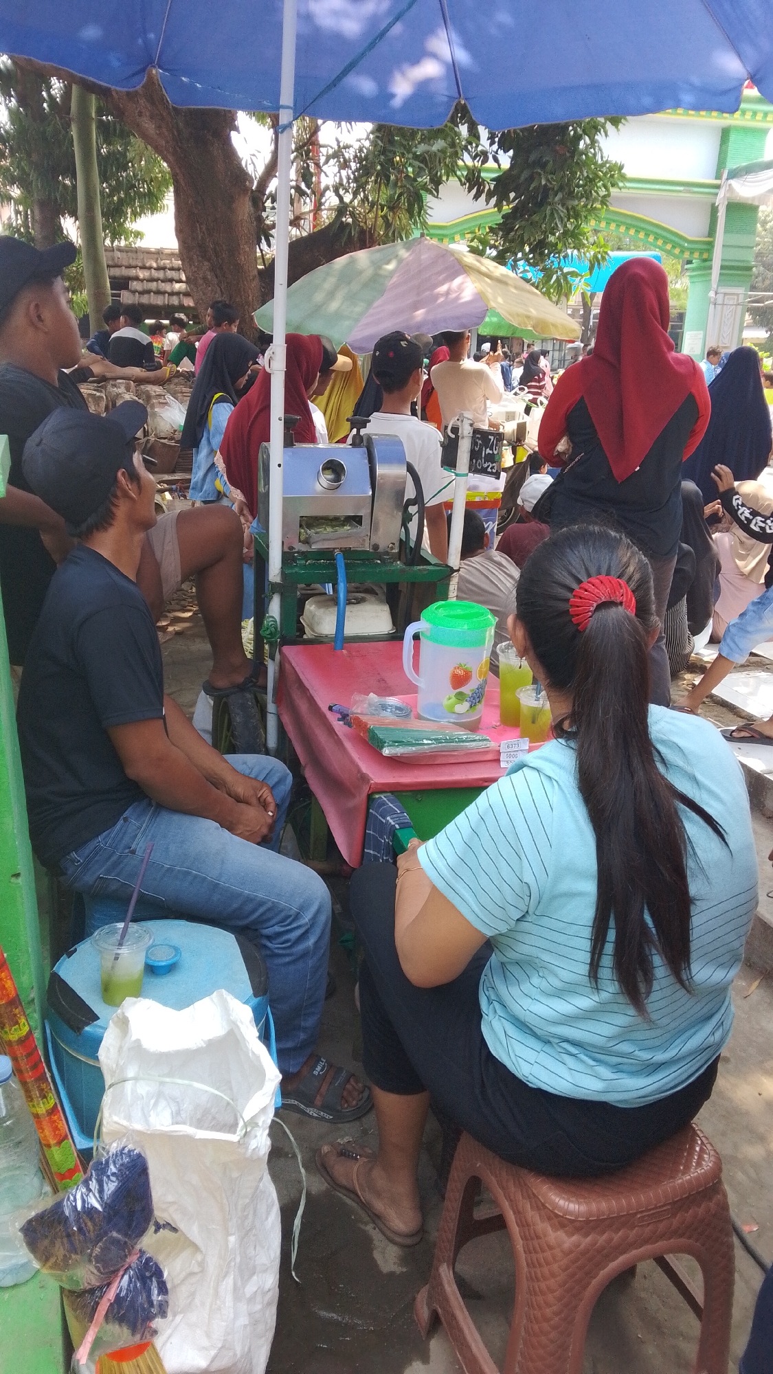 A group of people are gathered around a sugar cane juice stand under a blue umbrella. The stand is set up on a paved surface, likely a street or sidewalk. A sugar cane juicing machine is visible, along with a table covered with a red tablecloth. On the table, there are drinks, a pitcher, and green straws. Several people are seated and standing, some wearing traditional Indonesian clothing like headscarves. In the background, there is a green and white building with an arched entrance, characteristic of public buildings in Indonesia. The scene appears to be a bustling marketplace or a busy street corner in Mancar, Indonesia, where people are socializing and enjoying refreshments.