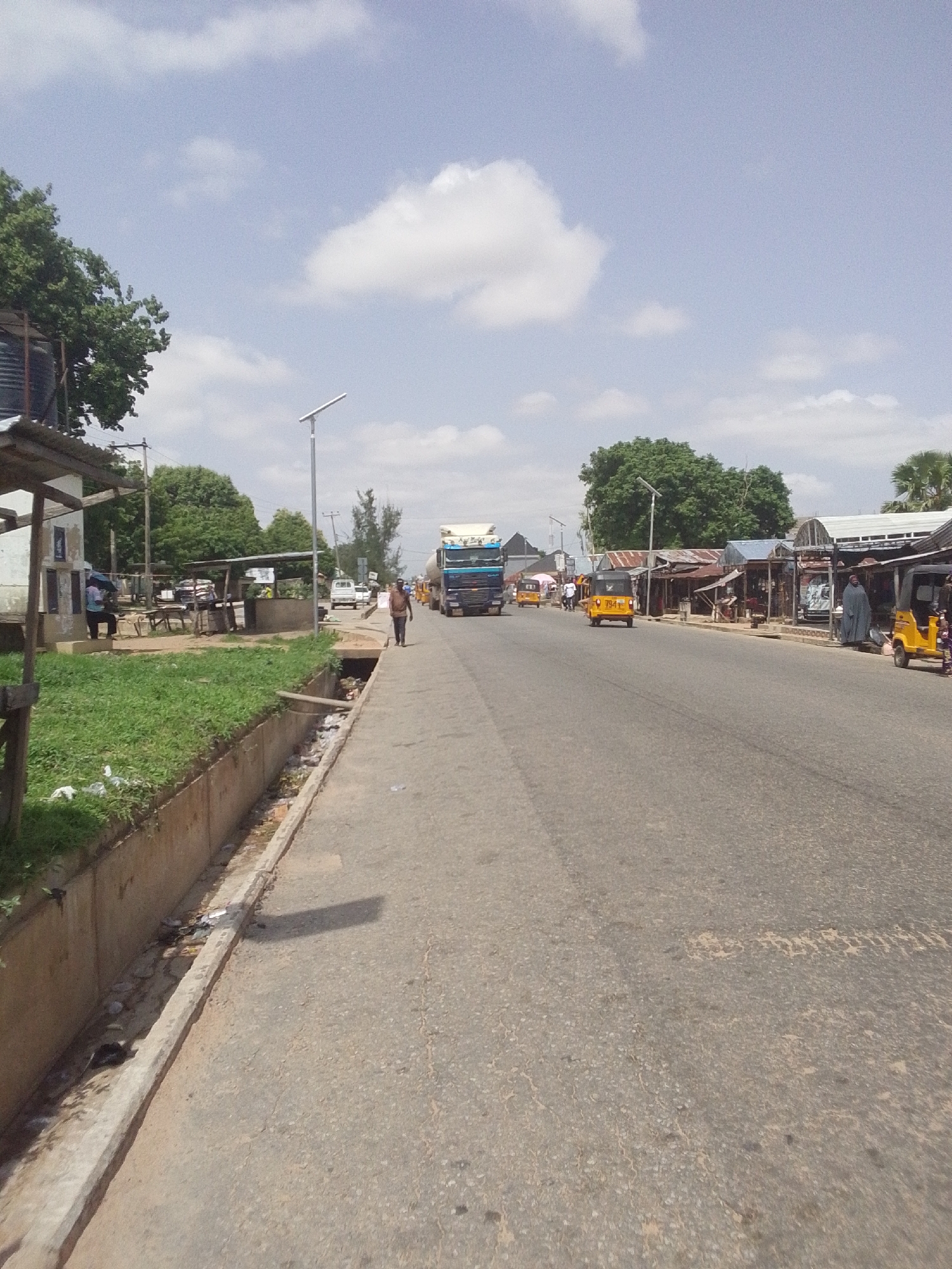 The image depicts a street scene in Jalingo, Nigeria, likely a commercial area. The road is paved and appears to be a main thoroughfare, with a concrete drainage channel running alongside it on the left. Green grass grows in patches by the drainage. Several buildings and market stalls line the right side of the road, many with corrugated metal roofs.
Vehicles present include a large blue truck in the background, several yellow auto-rickshaws (Keke Napeps), and a white car. People are visible, including a man walking on the sidewalk and another person standing near a market stall, holding an umbrella. The sky is partly cloudy with scattered white clouds against a pale blue background. The overall impression is of a bustling, everyday scene in a Nigerian city.