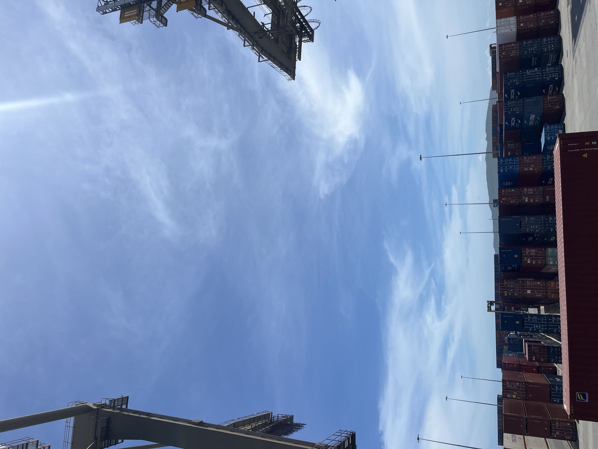 The image shows a view of a shipping port in Unknown, Unknown. The scene is dominated by a bright blue sky with scattered wispy clouds. In the top and bottom corners of the image are parts of large metal container cranes. To the right of the frame, stacks of shipping containers are piled high. Tall light poles are spaced between the containers.