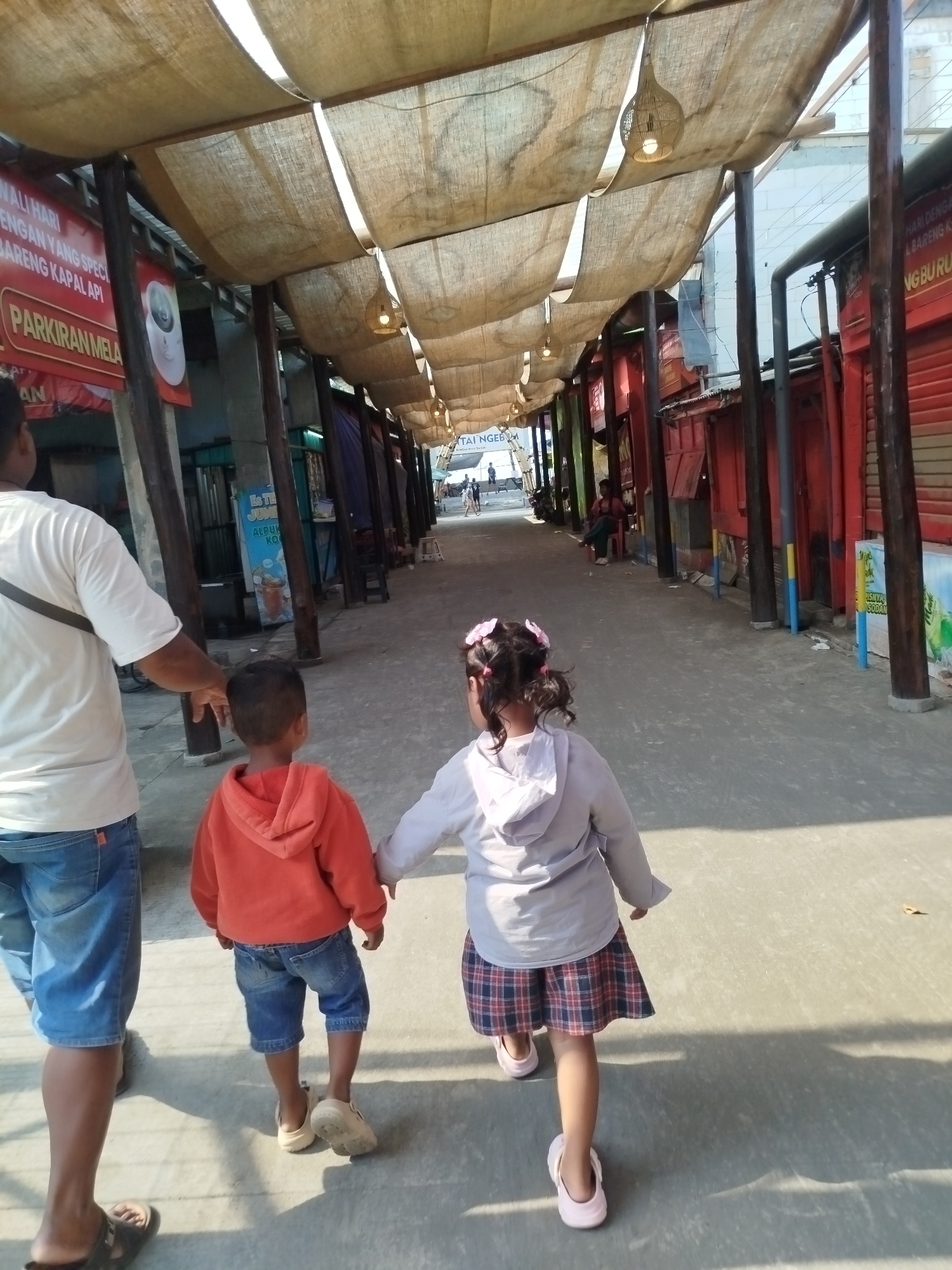 In the city of Mororejo, Indonesia, a family walks down a shaded pedestrian walkway. The walkway is covered by a light brown canvas canopy, supported by wooden beams, and lit by hanging woven lamps. On either side of the walkway, there are market stalls with red facades.
In the foreground, a man, seen from the waist down, wears a white t-shirt and blue denim shorts. He holds the hand of a young boy, who is wearing an orange hooded sweatshirt and blue denim shorts. The boy wears light-colored slip-on shoes. Next to the boy, a young girl, also seen from the back, wears a light grey hooded jacket and a plaid skirt. She has pink bows in her hair. The girl wears pink slip-on shoes. The family is walking away from the camera down the walkway.
In the background, other people can be seen walking and sitting at the market stalls. The ground of the walkway is paved with concrete. The overall scene depicts a lively, outdoor market environment on a sunny day.
