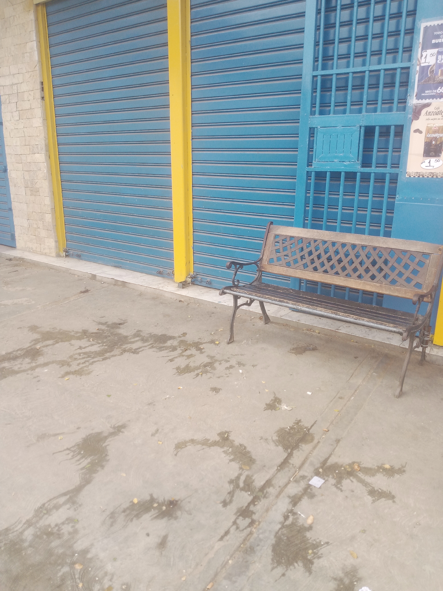 The image depicts a street scene with a blue roll-up shutter and a blue metal gate in the background. To the left of the shutter is a wall constructed with light-colored bricks. In front of the shutter and gate, a weathered wooden bench with metal legs is placed on a concrete surface. The concrete appears damp and shows some scattered leaves and debris. A portion of a poster is visible on the blue gate, with text in Spanish including "Anzoátegui" and prices indicated. The overall impression is an outdoor commercial or storefront area in an urban setting, specifically identified as Santa Ana, Venezuela. There are no people or discernible actions taking place in the image.