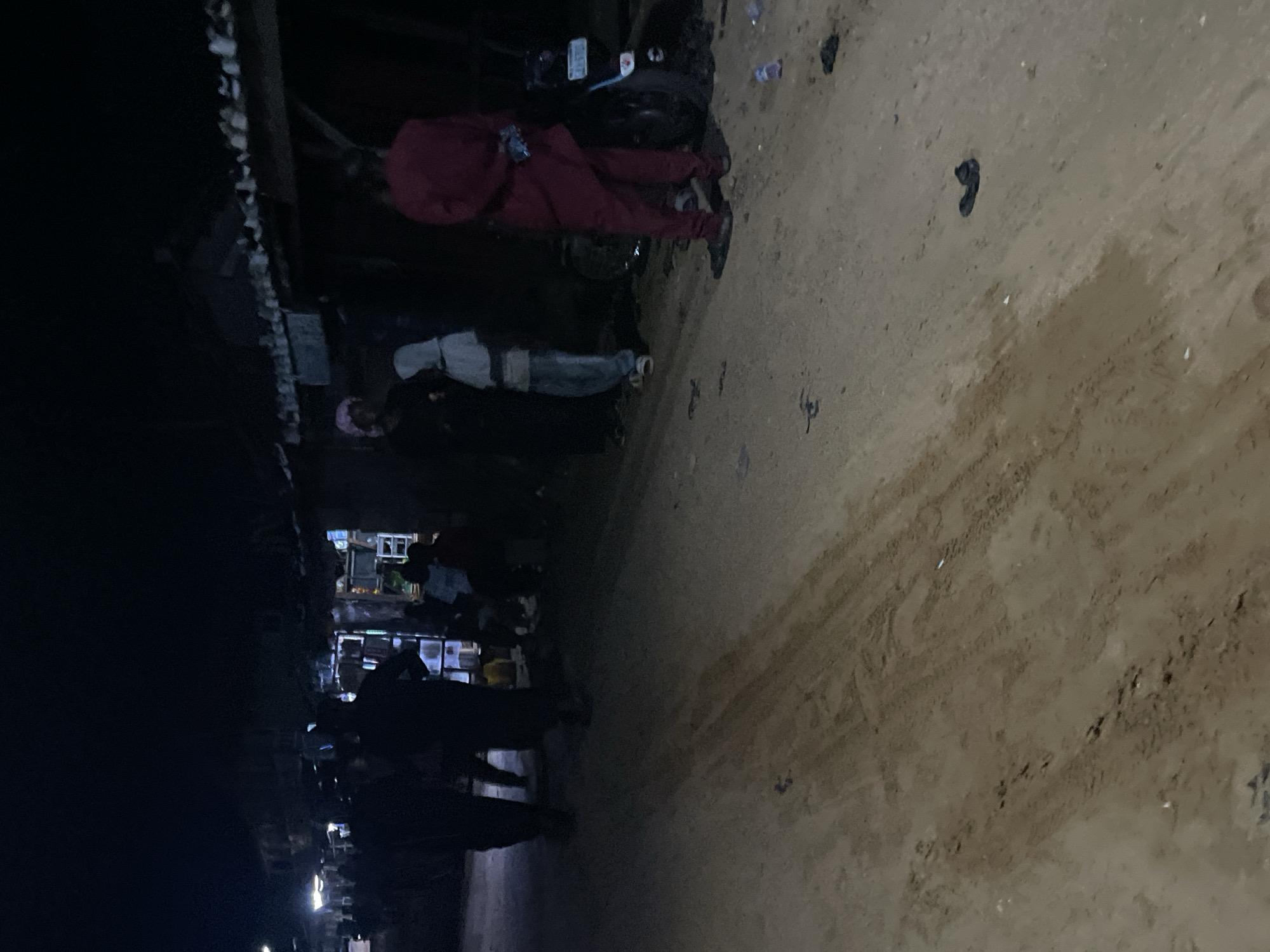 The image depicts a street scene at night in Lokoja, Nigeria, with several individuals gathered. The ground is unpaved and appears to be a dirt road. Buildings line the side of the street, some with illuminated storefronts. A motorcycle is parked on the right side of the image. People are standing and walking, with some appearing to be interacting. The lighting suggests artificial illumination from the buildings and possibly some streetlights. The overall atmosphere is that of a busy, albeit dimly lit, public space.