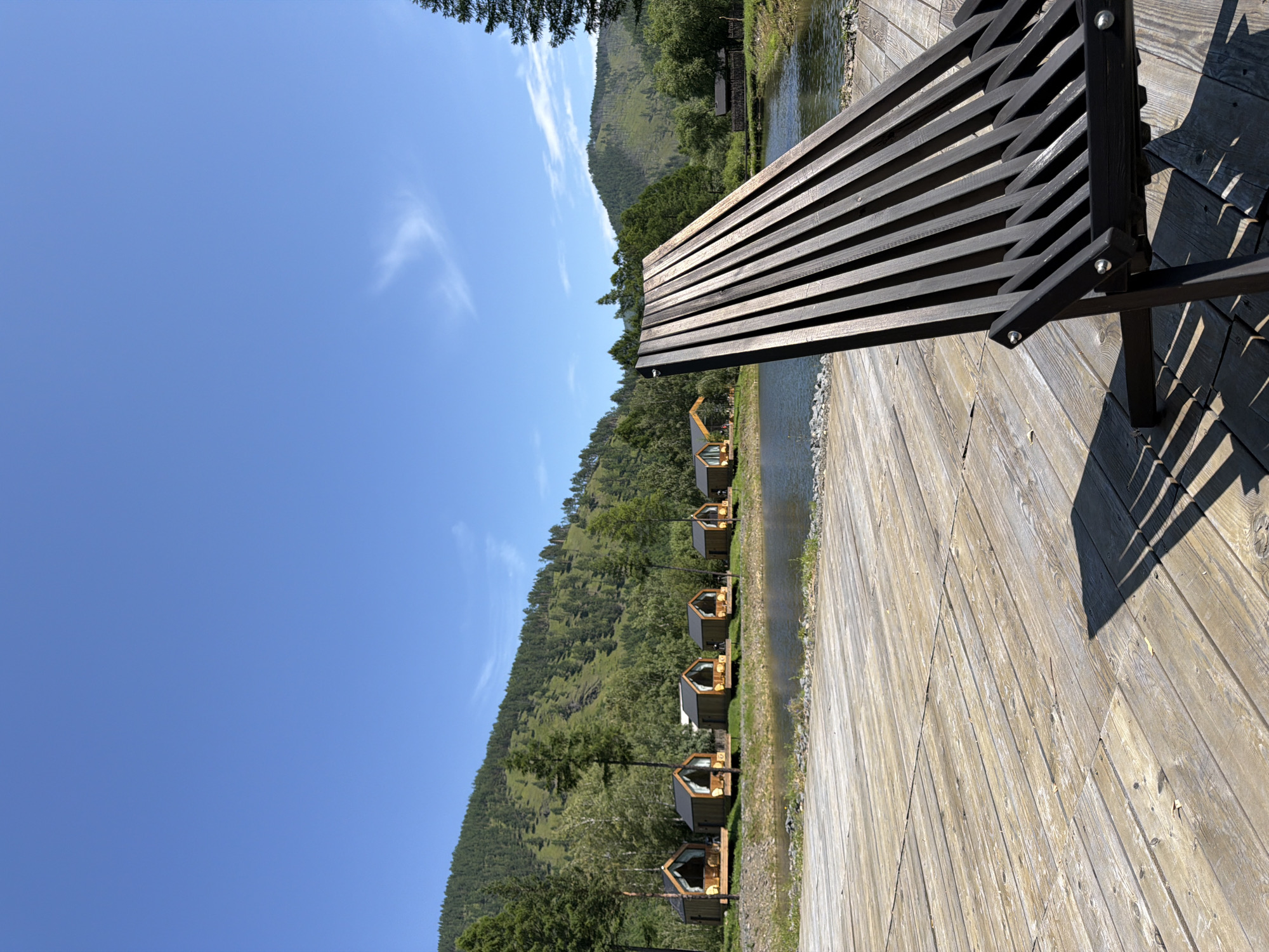 A scenic view of a resort area with small wooden cabins situated along the bank of a tranquil lake, with a lush, green, tree-covered mountainside in the background. The foreground is dominated by a wooden deck, upon which rests a dark wooden lounger chair, casting a patterned shadow across the weathered planks. The sky is a clear, vibrant blue with a few wispy white clouds. The cabins, each with a triangular roof and large glass front, appear unoccupied. The water of the lake is calm and reflects the surrounding landscape. The overall impression is one of peaceful, natural beauty, likely a tourist destination.