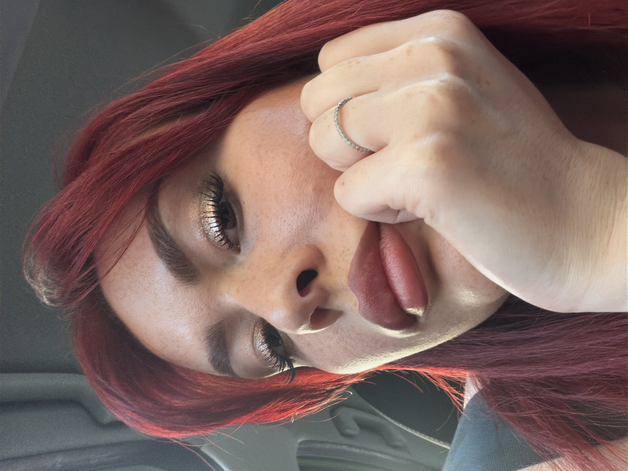 The media file shows a close-up, angled selfie of a young woman with vibrant red hair. She is wearing makeup, including shimmery eyeshadow and lipstick, and has a delicate ring on her right hand, which is resting on her cheek. Her tongue is slightly protruding, adding a playful element to the expression. The lighting suggests it might be daytime, possibly indoors given the smooth background. There are no visible text elements in the image. The setting is not clearly defined, but the presence of what appears to be car upholstery in the background might indicate she is in a vehicle. The overall mood is casual and perhaps a bit flirty.