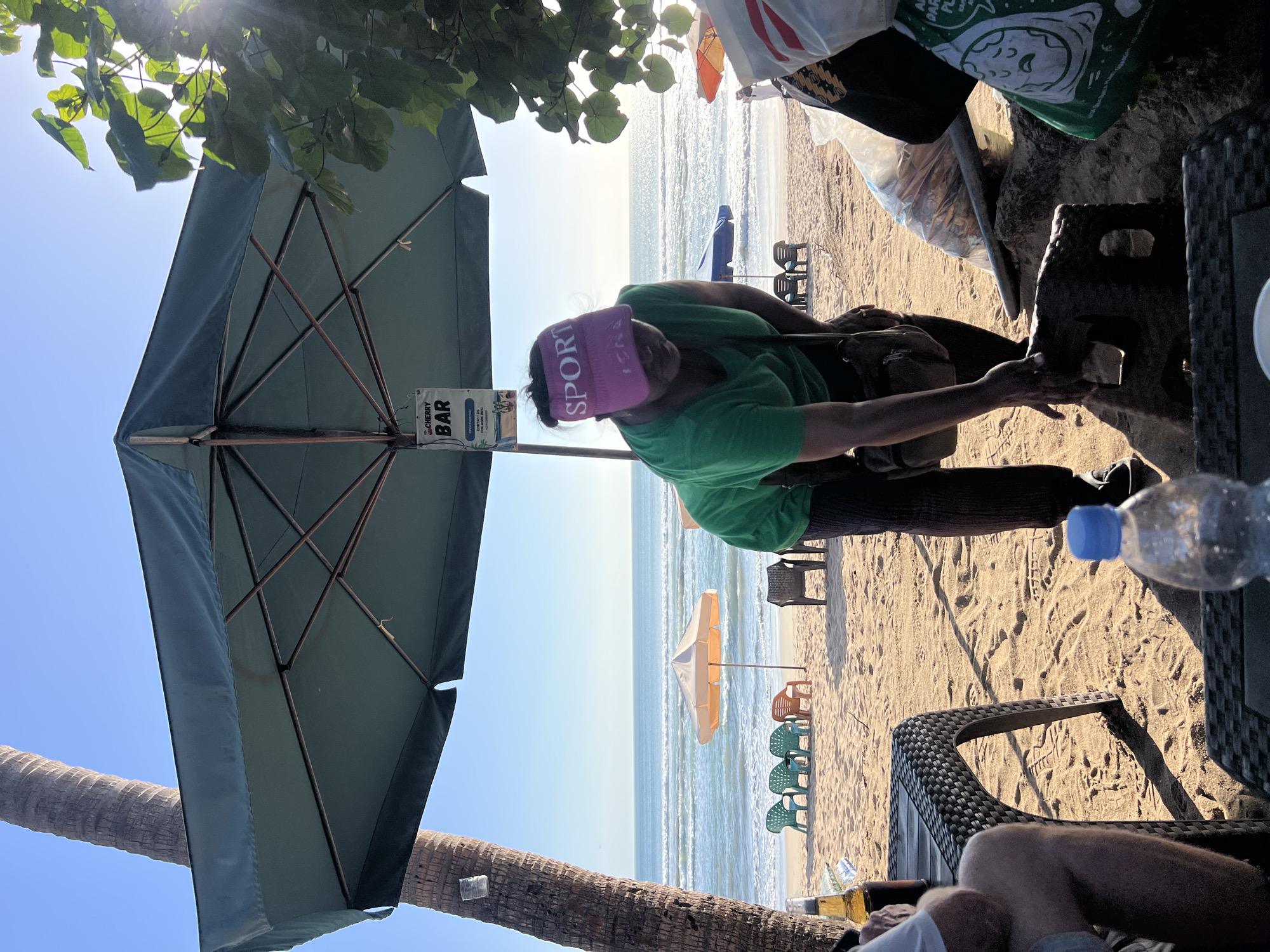 The image depicts a beach scene in Kuta, Indonesia, likely in the morning or late afternoon due to the low angle of the sun casting long shadows. A person wearing a green shirt and a pink "SPORT" visor is bent over, interacting with an object on the sand. They have a bag slung across their shoulder. In the foreground, a plastic water bottle sits on a woven table. To the left, a person wearing a white shirt and light-colored shorts is partially visible, with a bottle of beer on the table. Behind them, a large green umbrella provides shade. Further back, the ocean stretches to the horizon with gentle waves. Beach chairs are arranged on the sand, some in clusters, and a small orange umbrella is visible near the water. A sign hanging from the large umbrella reads "CHERRY BAR". The overall environment is a tropical beach with palm trees and vegetation.
