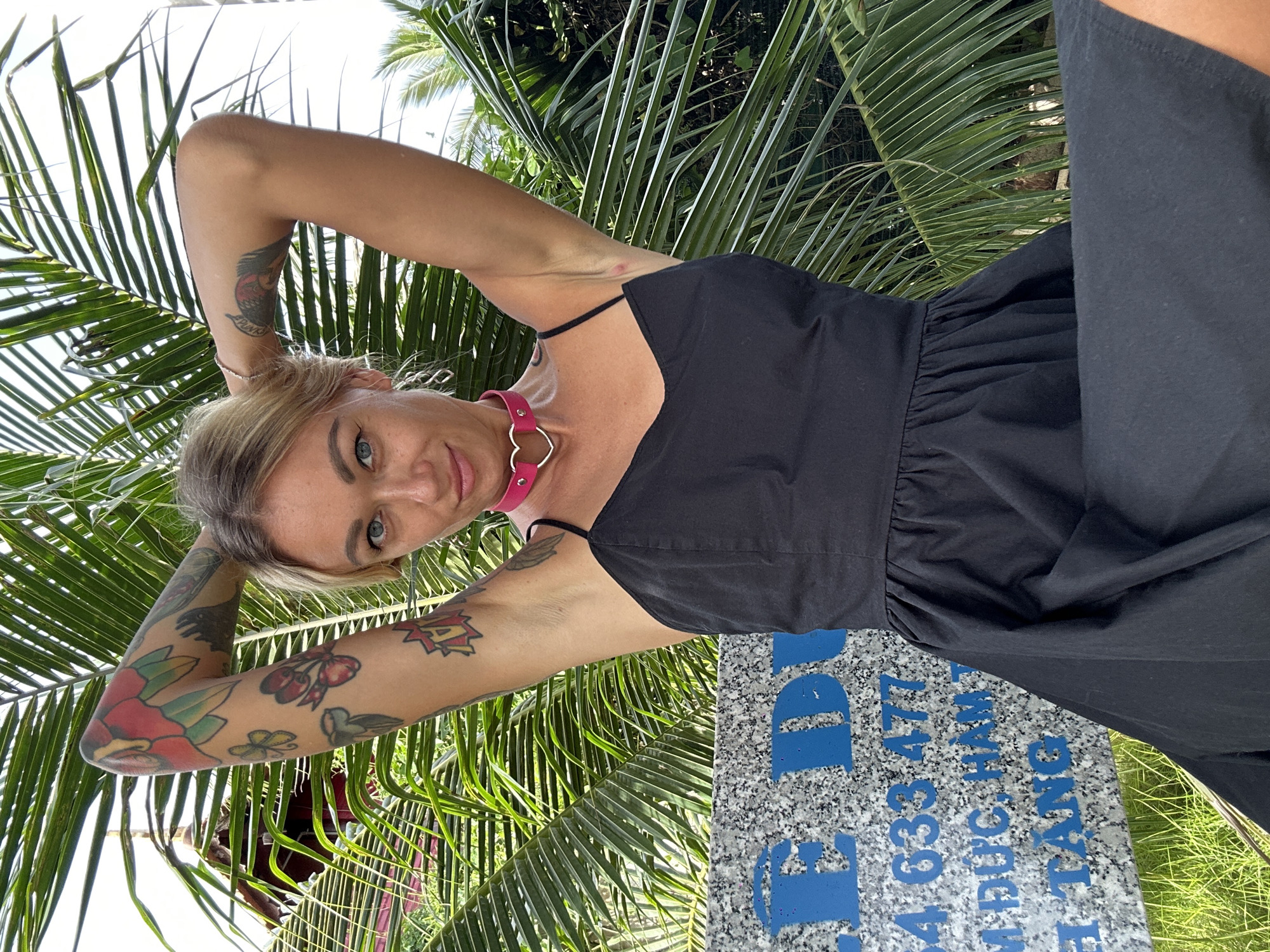 A woman with blonde hair, blue eyes, and visible tattoos on her arms is posing outdoors, with her hands behind her head. She is wearing a black dress and a pink choker with a heart pendant. Behind her are large palm fronds and a sign with blue lettering, which appears to be in Vietnamese and contains numbers. The sign has text that reads "4 633 477" and "M DUC HAM" and "TANG". This scene is located in Mũi Né, Vietnam.