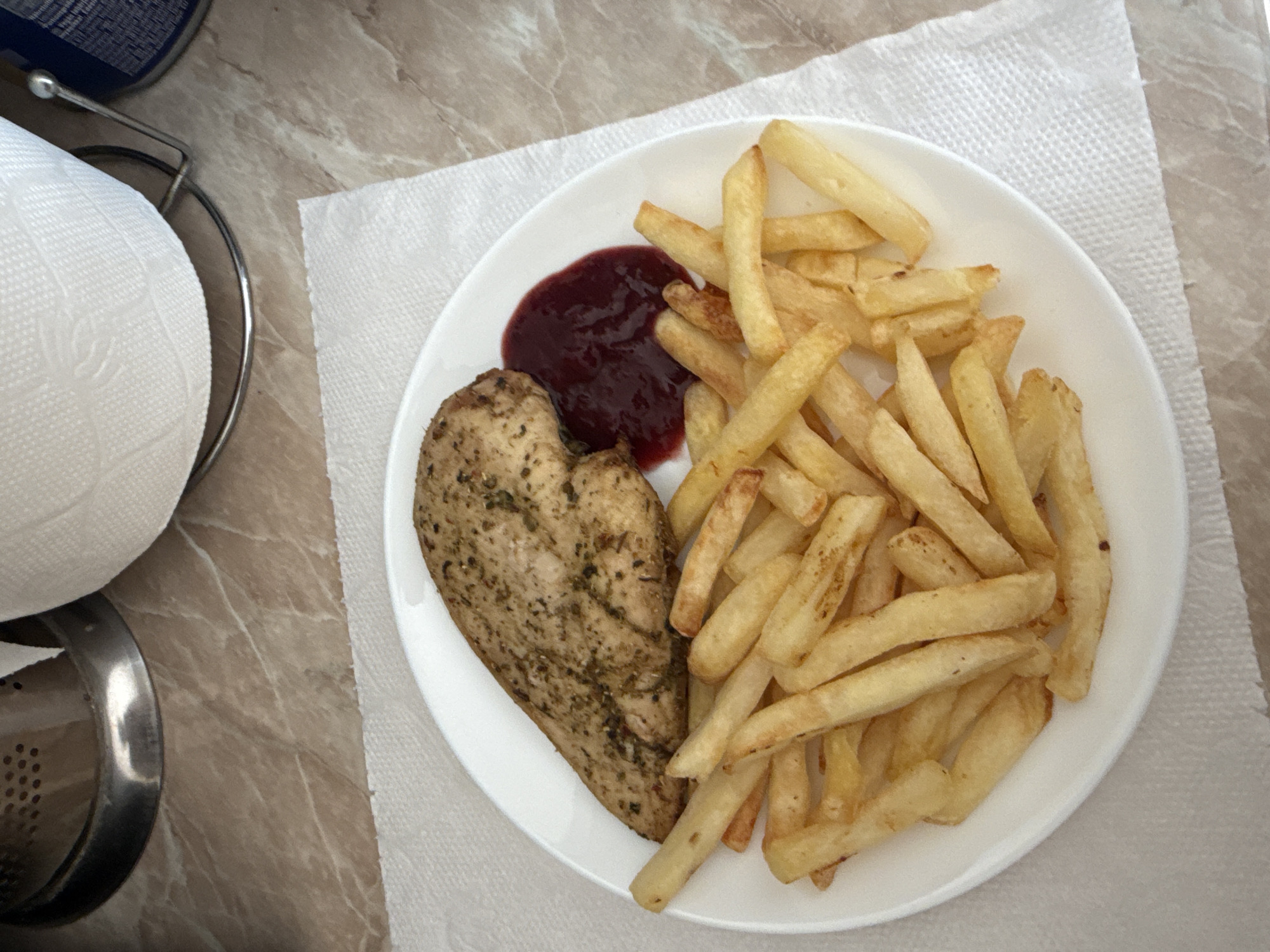 A close-up, overhead shot captures a white plate filled with food, resting on a white paper napkin. The plate contains a seasoned, cooked chicken breast and a generous portion of french fries. To the right of the chicken breast, there is a small dollop of dark red sauce, possibly cranberry or berry sauce. The background shows a marbled countertop, a roll of paper towels on a stand, and a part of a metal object. The lighting suggests an indoor setting, possibly a kitchen or dining area. The image does not provide any specific clues or context that would indicate its location in Tbilisi, Georgia, or any specific city.