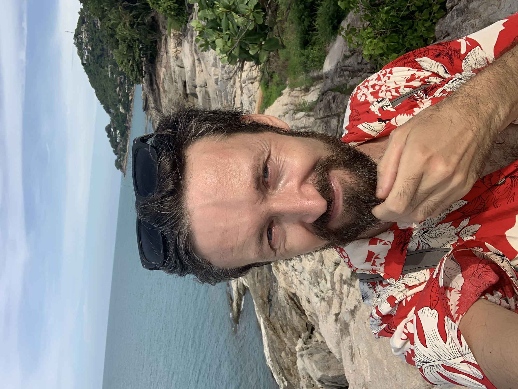 A man with a beard and sunglasses perched on his head is taking a selfie. He is wearing a red and white floral shirt. The background shows a rocky coastline with the ocean and a lush green hillside dotted with buildings. The scene appears to be a tourist destination, possibly in Baan Chaweng Noi, Thailand, given the tropical vegetation and coastal landscape. The man is touching his beard with his fingers. The lighting suggests it is daytime.