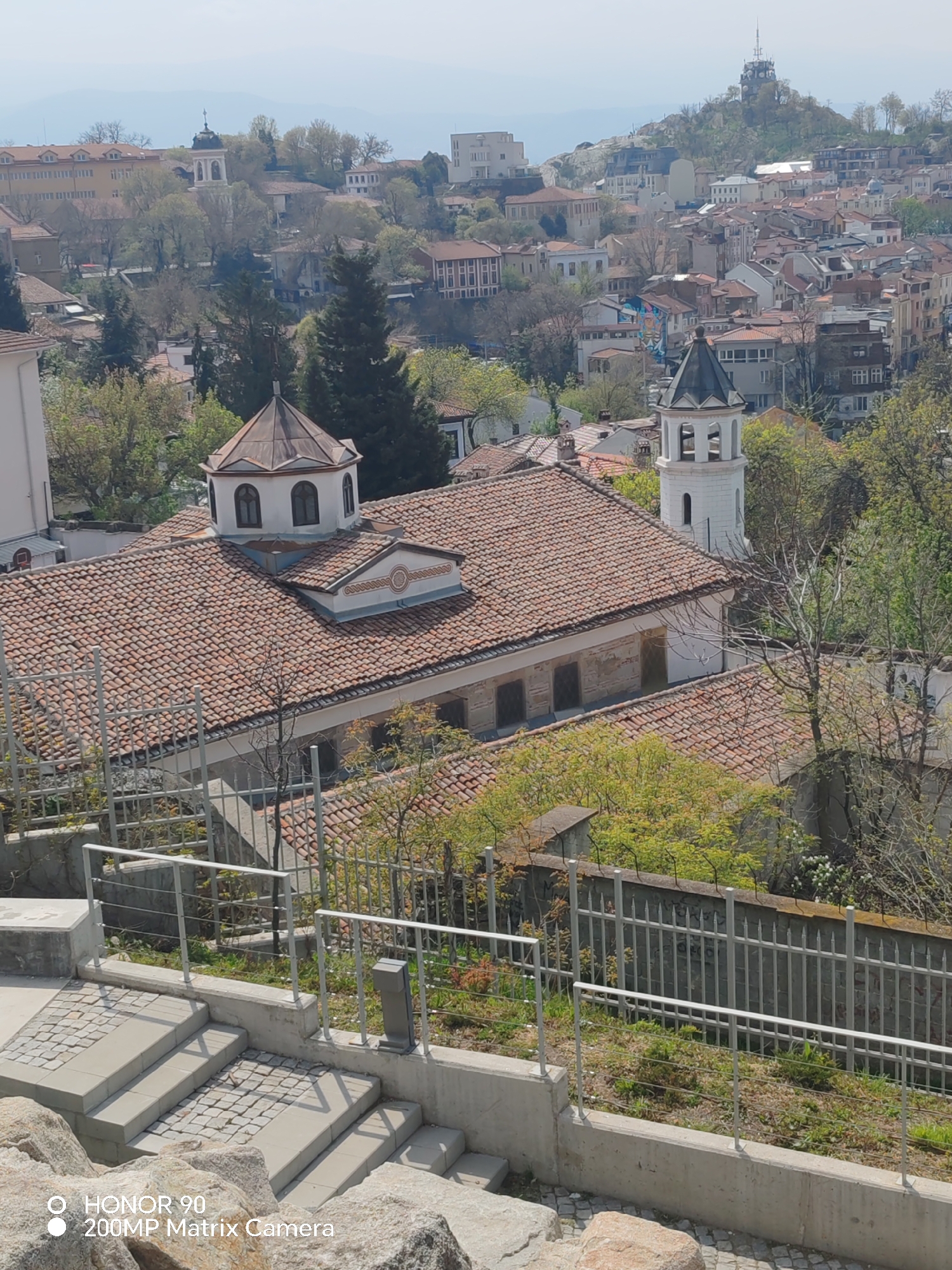 This is an eye-level outdoor shot taken in the city of Plovdiv, Bulgaria. The perspective is from an elevated position looking out over the city. In the foreground are stone steps leading down, partially blocked by a metal railing, and a low concrete wall.
The primary subject is a church with a clay-tile roof, a central dome, and a white bell tower. To the left of the frame, a segment of a white building is visible. Trees are interspersed around the building and city landscape.
The middle ground features a dense urban area composed of buildings, houses, and other structures of varying sizes and architectural styles. The buildings are arranged closely together.
In the distant background, a hill with a tower is visible. The sky is overcast.