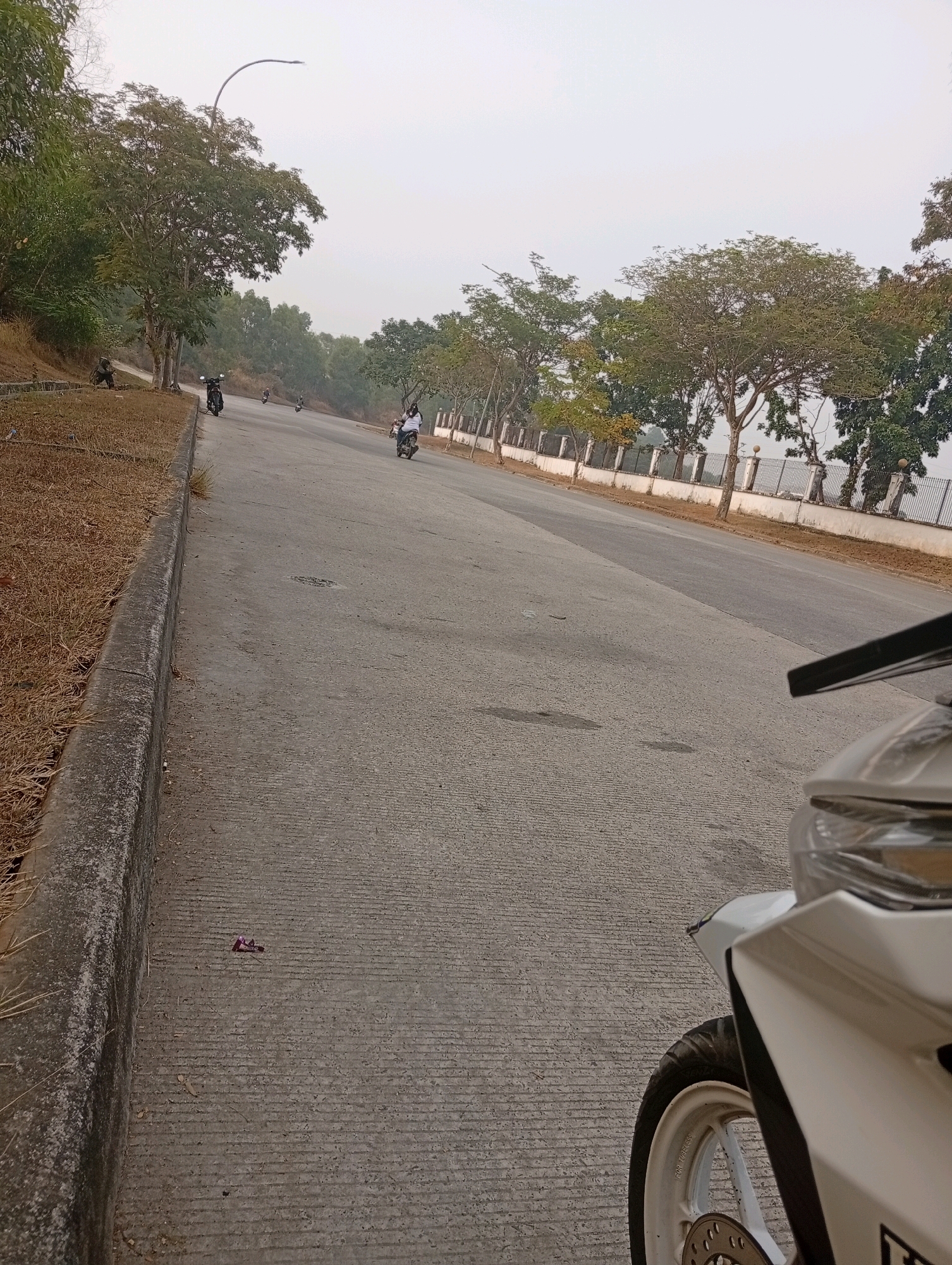 The image depicts a street scene in Sukamahi, Indonesia. The road is paved with concrete and marked with visible tire tracks. On the left side of the road, there is a grassy embankment with dry vegetation and a concrete curb. Several trees line the right side of the road, behind a decorative fence. A lamppost with a distinctive curved design is visible in the background.
Several motorcycles are present on the road. One motorcycle, partially visible in the foreground on the right, is white with black accents and a white wheel. Three other motorcycles are seen further down the road, each with a rider. One rider is wearing a white shirt and black pants, another is wearing a dark shirt and pants, and a third is wearing a light-colored shirt and dark pants. The overall atmosphere appears to be hazy, suggesting a morning or evening setting, or possibly atmospheric conditions. The scene is devoid of any immediate signs of activity beyond the presence of the motorcycles and riders.