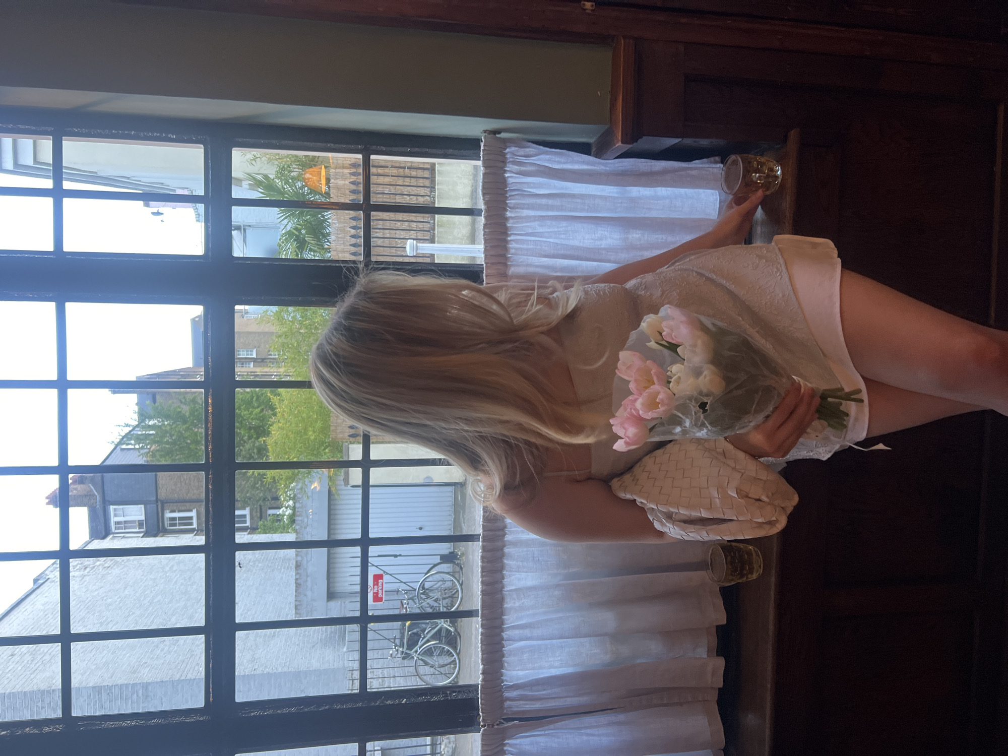 A young woman with long, blond hair sits at a window with a grid pattern. She wears a light-colored dress. In her hands, she holds a bouquet of pink and white tulips and a cream-colored woven handbag. She appears to be inside a building. Next to her is a glass of liquid. Outside the window is a street scene with bicycles and residential buildings, suggesting the location is in London, United Kingdom.
