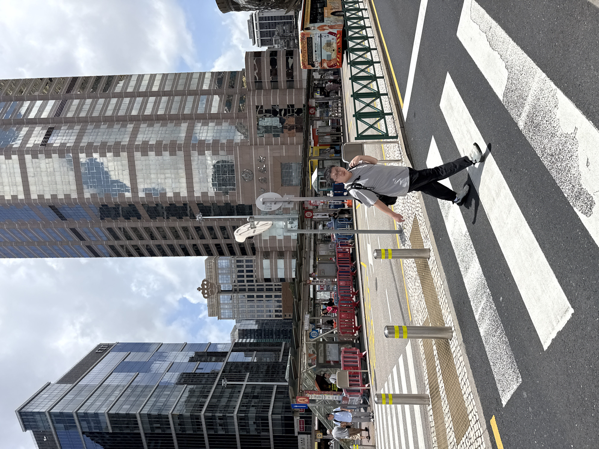 The image captures a street scene in Macau, China, dominated by modern high-rise buildings. In the foreground, a young man wearing glasses, a grey t-shirt, and black pants is crossing a pedestrian zebra crossing. He is carrying a black backpack over his right shoulder and appears to be looking towards the right. The zebra crossing is marked with white stripes on dark asphalt, bordered by a tactile paving surface. Several bollards with yellow stripes are positioned along the crossing.
In the mid-ground, a bus stop area is visible with people waiting. Red and white barriers cordon off the waiting area. A bus with an advertisement on its side, featuring Chinese characters and images, is parked at the stop. Behind the bus stop, several tall buildings rise, one with a distinctive crown-like architectural element at its top. The buildings have reflective glass facades, showing a cloudy sky. A green metal fence runs along the side of the road.
The background is filled with contemporary urban architecture, including glass-fronted office buildings and other multi-story structures. The sky is partly cloudy. The overall environment suggests a busy urban setting in Macau.