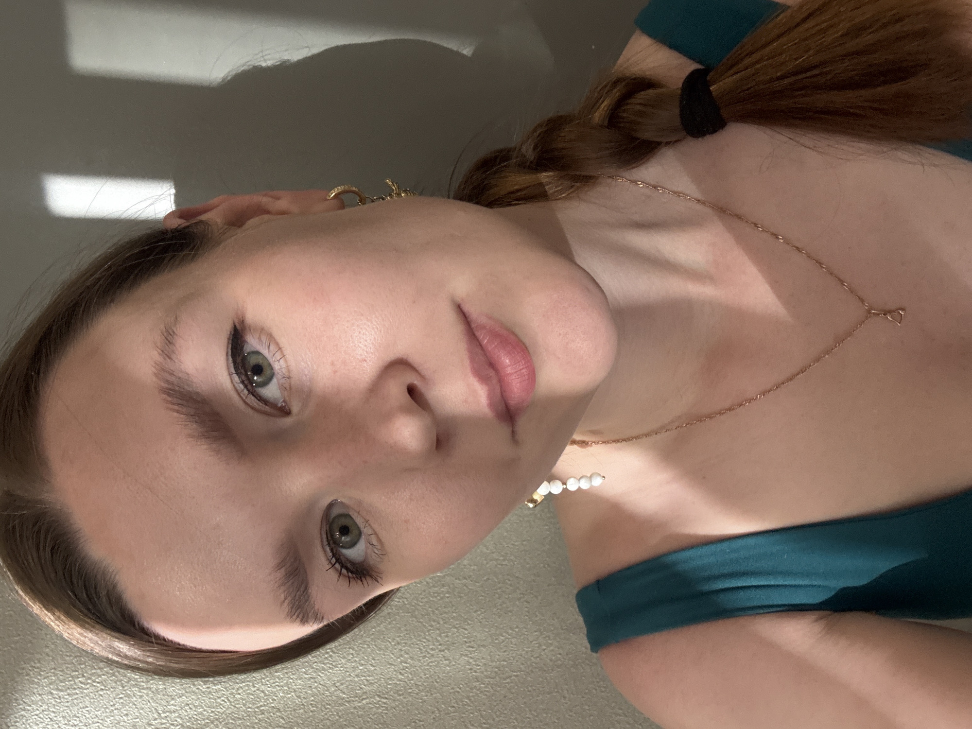 A close-up, angled portrait features a young woman looking directly at the camera with a calm, neutral expression. Her head is tilted sharply to her right, and the photo appears to be taken from a low vantage point, possibly a selfie. She has light green eyes, defined eyelashes, and dark, full eyebrows. Her lips are a natural pink color and slightly parted.
Her long, reddish-brown hair is styled in a single braid, secured with a black elastic band, and rests over her left shoulder. She is wearing a dark teal or green top, possibly off-the-shoulder or strapless. Accessories include a gold hoop earring visible on her right ear, a delicate gold chain necklace with a small, simple pendant around her neck, and a white pearl bracelet on her left wrist.
The indoor setting shows a light-colored wall in the background, illuminated by strong, angular shadows cast by natural light, suggesting it is daytime and sunlight is entering through a window. No visible text is present in the image. This media file was taken in Izhevsk, Russia.