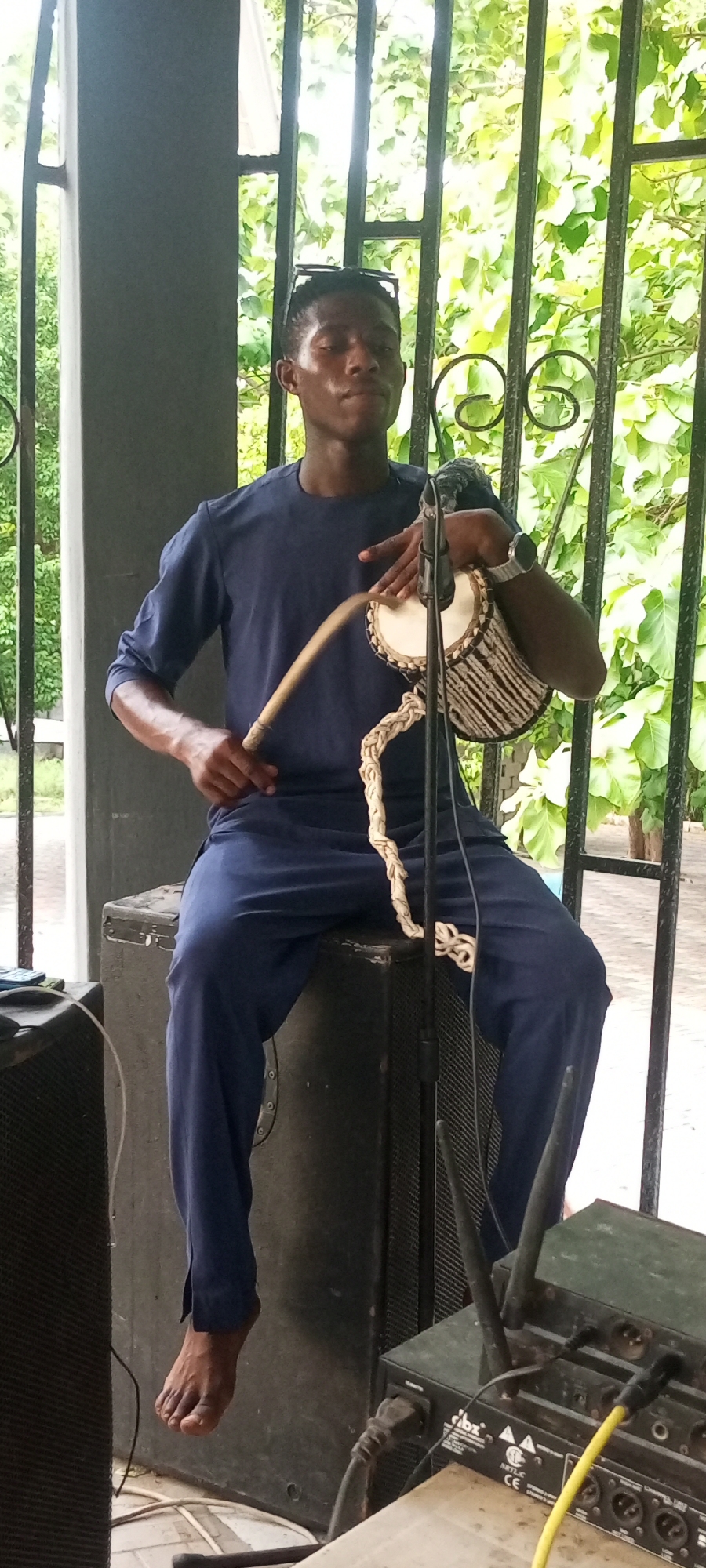 The image depicts a man seated on a dark-colored, presumably metal, speaker box, playing a small drum with a wooden stick. The man, appearing to be of African descent, is wearing a blue outfit, and sunglasses are resting on his head. He is barefoot, and a microphone is positioned in front of him. The drum has a light-colored membrane with a darker patterned body and a braided rope attached. To the right of the man, an audio mixer with multiple inputs, antenna, and power connections is visible. The backdrop features an ornamental metal fence, and beyond it, greenery indicative of a park or garden setting. The location is Unknown, Unknown.