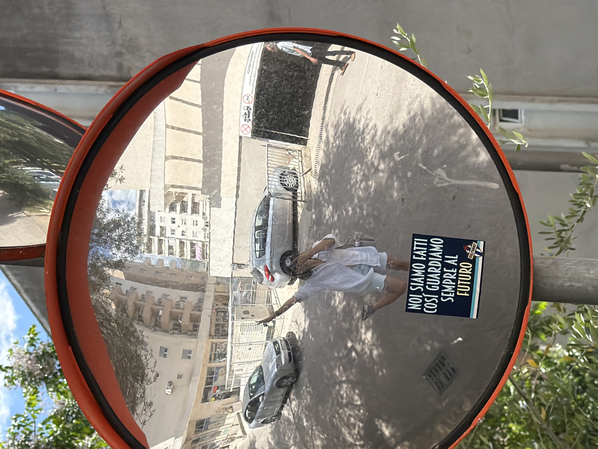 The image is a street scene reflected in a convex safety mirror. The mirror is round, with an orange frame and a black edge. In the reflection, two people are visible, one of whom appears to be taking a photo of themselves and the mirror. Two cars are also reflected, parked on a street next to buildings and a parking garage. A sticker on the mirror reads "NOI SIAMO FATTI COSI GUARDIAMO SEMPRE AL FUTURO" (WE ARE MADE LIKE THIS WE ALWAYS LOOK TO THE FUTURE). The overall location is Budva, Montenegro, as suggested by the environment and the local language of the sticker. The mirror is mounted on a pole, and parts of trees are visible in the periphery, suggesting it's located on a street with vegetation.