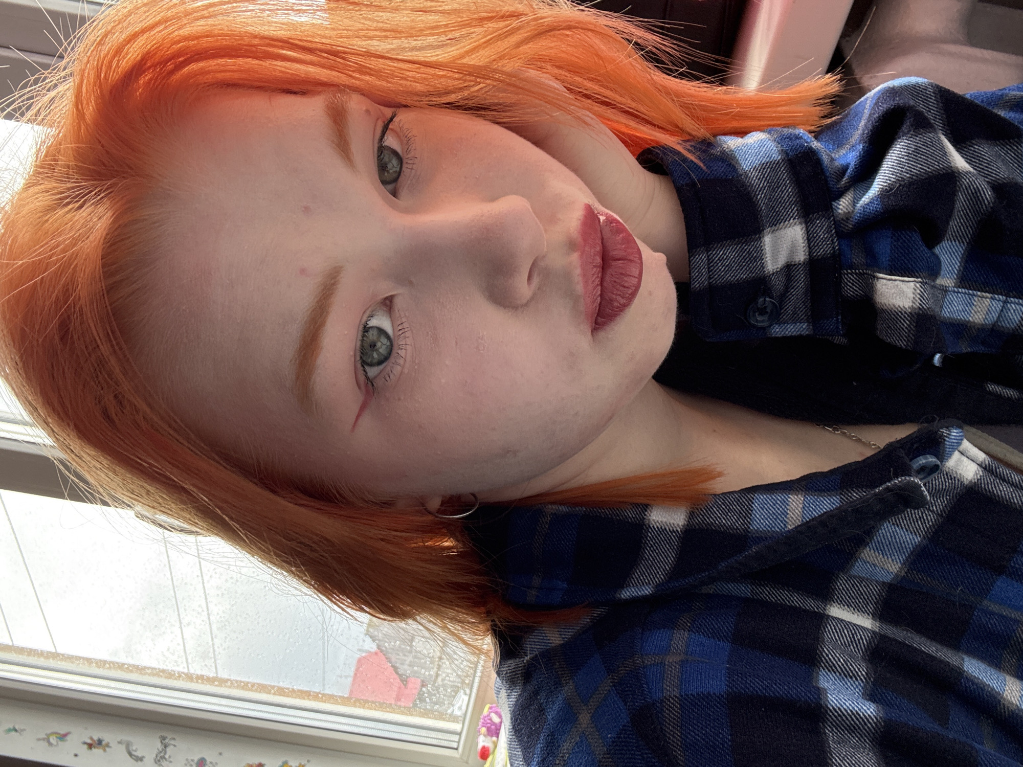 A medium shot captures a young woman with light skin and vibrant orange hair. She wears a blue and black plaid shirt. Her green eyes gaze directly at the viewer. Her face is angled slightly to the side, and her lips are pursed. A thin silver hoop earring is visible. A window is partially visible in the background. The location is Unknown, Unknown.