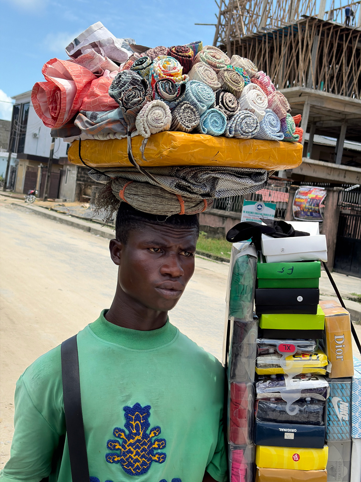 A young Black man stands on a street in Igboefon, Nigeria, carrying a large load of goods on his head. The goods are stacked in multiple layers, with the top layer consisting of neatly rolled fabric, many of which appear to be patterned. Below this are other items wrapped in yellow packaging. The man is wearing a green long-sleeved shirt with a blue and yellow embroidered design on the chest. He has a dark sling bag across his shoulder. To his right, there is a vertical stack of boxes, some of which are labeled with sizes such as "XL" and "XXL," and brand names like "Dior" and "Nike." In the background, there is a partially constructed building with scaffolding made of wood. The sky is bright and appears to be daytime. The street is paved with asphalt, and there are buildings and a motorcycle visible in the distance.