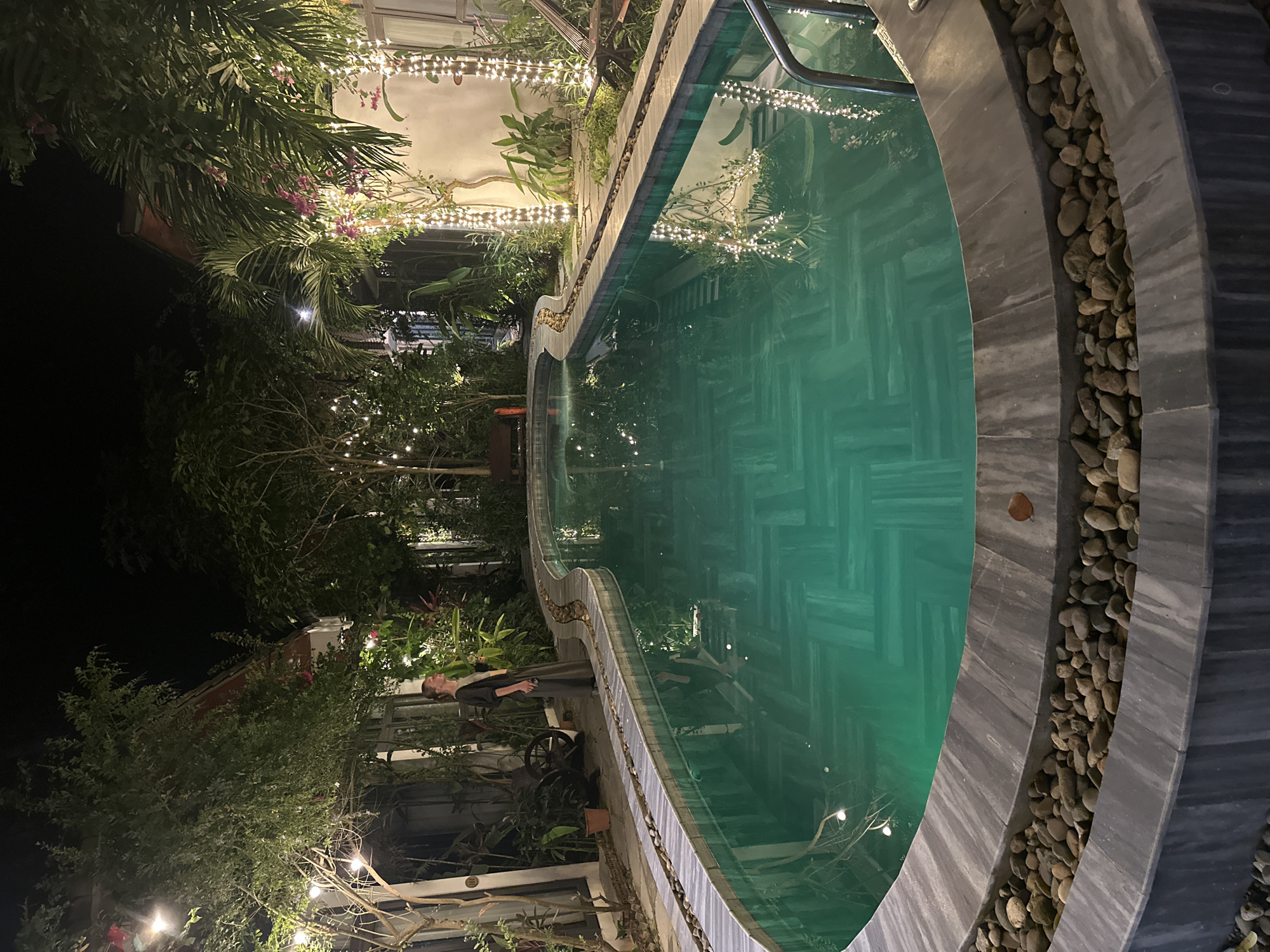 A medium shot captures a serene, curved swimming pool at night, illuminated by numerous small, white string lights and ambient outdoor lighting. The pool water is a vibrant turquoise, revealing a patterned tile bottom. A woman stands to the left of the pool, facing away from the camera, her silhouette outlined by the lights. She is wearing a long, dark skirt and a lighter-colored top. The pool area is surrounded by lush vegetation, including trees and tropical plants, with some lights intertwined with branches. Buildings with red-tiled roofs are visible in the background, partially obscured by the foliage. The pool deck is finished with stone tiles and a border of small, smooth pebbles. The overall impression is one of a tranquil, tropical night scene. The image appears to be taken in the city of Nam Hoa Lư Ward, Vietnam.
