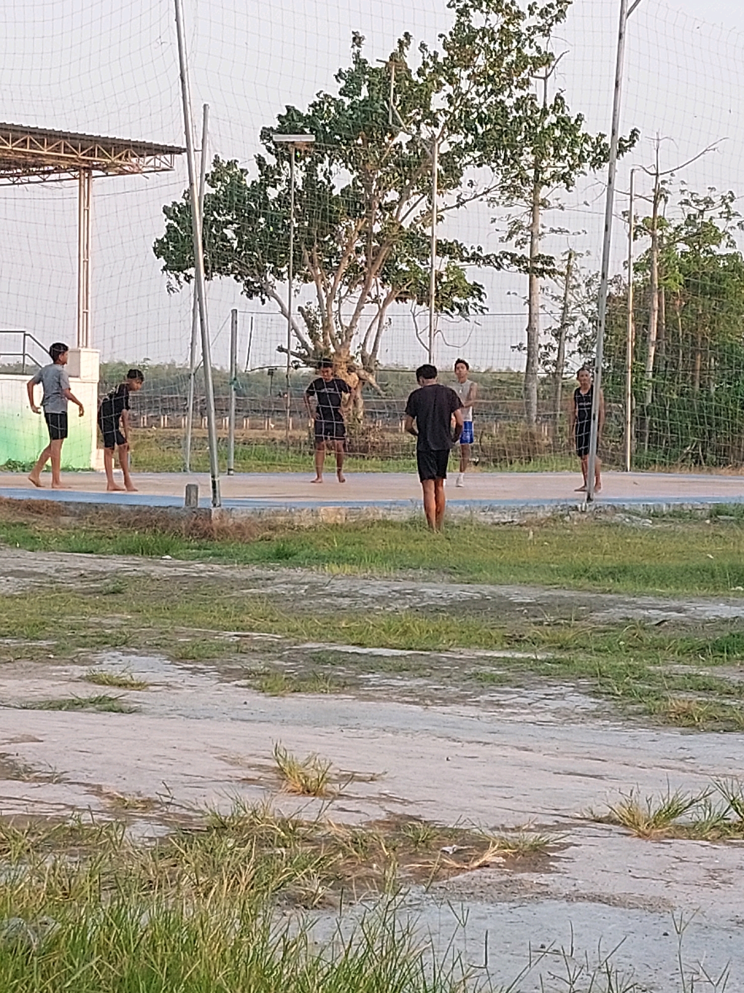 In the city of Canga'an, Indonesia, a group of young men are gathered on an outdoor court, possibly for a game of volleyball. The court is marked with blue and white lines, and a net stretches across it, supported by metal poles. Several trees with green foliage surround the court, and a wire fence encloses the area. In the foreground, dry grass and patchy dirt cover the ground. One individual in the foreground is facing away from the camera, dressed in a black t-shirt and shorts, while another in a grey t-shirt and dark shorts walks to the left. In the background, two other individuals are visible on the court. The sky above is a pale, overcast color.
