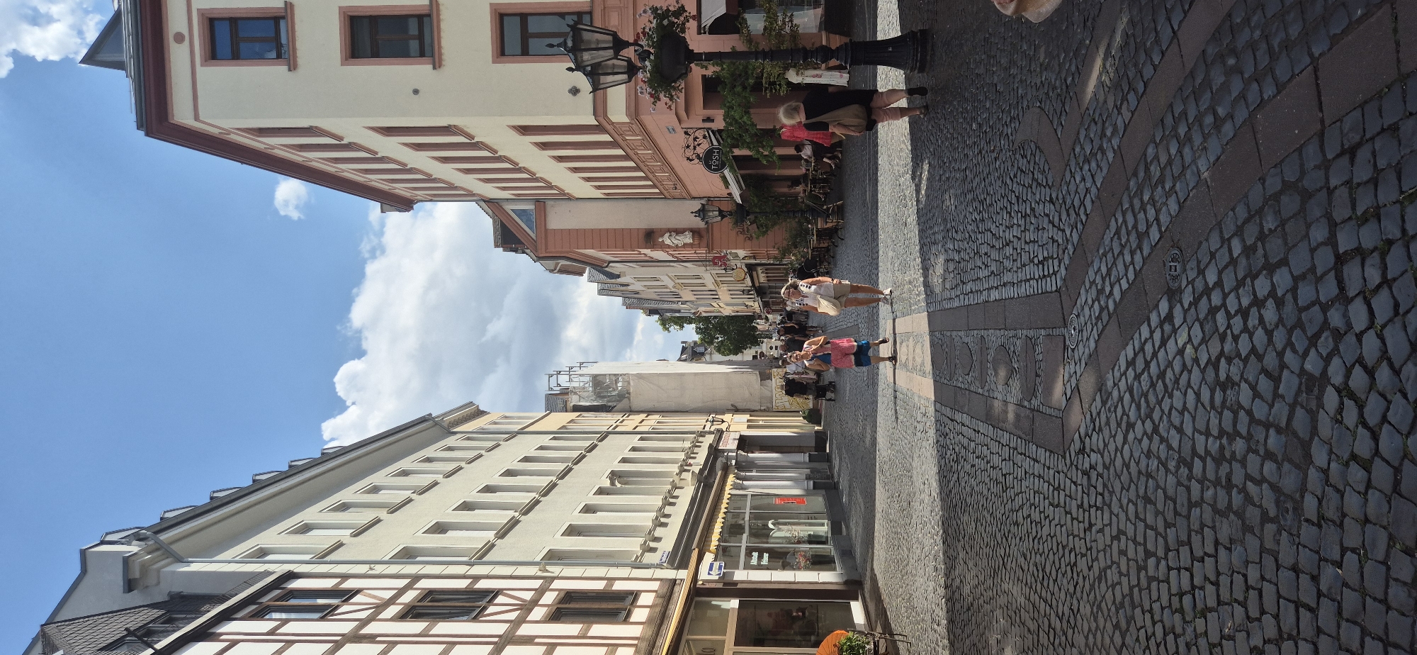 The image shows a cobblestone street in Mainz, Germany, lined with historic buildings. The sun is shining, casting shadows on the street. Several people are walking in the street, some in groups and others individually. One woman in the foreground is wearing a pink and blue dress and walking towards the camera. To her right, another woman is wearing a white top and shorts and walking away from the camera. There are shops and cafes on either side of the street, with outdoor seating areas visible. A decorative streetlamp with flowers is also visible. The architecture suggests a historic European city.