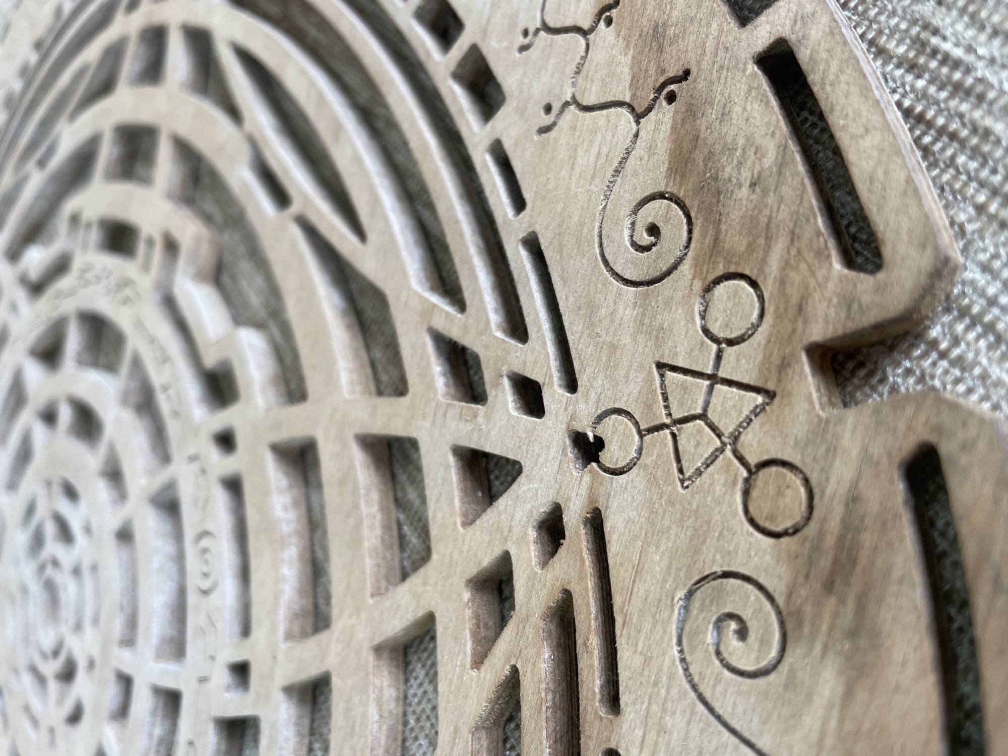 The image shows a close-up of a wooden object with intricate carvings and cutouts. The object appears to be a circular or semi-circular decorative piece, possibly a lid or a wall hanging. The surface of the wood is light brown and shows some grain texture. The carvings include geometric patterns, spiral designs, and what appear to be symbolic figures. One symbol, centrally located in the lower part of the image, resembles a stylized stick figure with a circle for a head and an outstretched arm, holding what looks like a ring. Another figure to the right appears more abstract, with delicate lines and dots. The background is a textured fabric, possibly burlap, which is a neutral beige color. The lighting creates shadows that emphasize the depth and detail of the carvings. The image does not provide any information about the city of Bhagsu, India, or any people or actions occurring within it.