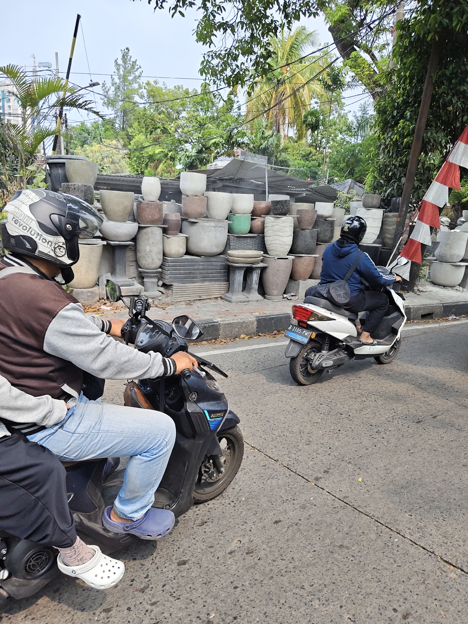 The image depicts a street scene in South Jakarta, Indonesia, likely near a roadside vendor selling pots. In the foreground, two people are riding a black scooter. The driver, wearing a black helmet with white accents and a brown and grey jacket, is in control of the scooter. The passenger behind them is wearing light blue jeans and a grey hoodie. Their feet are visible, with the passenger wearing white Crocs and a patterned sock, and the driver wearing blue Crocs.
The scooter has a blue license plate with white lettering that reads "B 3105 PWG".
To the left and behind the scooters, a large display of various sizes and styles of pots is arranged. These range from simple grey concrete pots to more decorative textured ones, some appearing to be made of clay or a similar material. The pots are stacked on shelves and directly on the ground.
In the background, lush green trees and palm fronds are visible, indicating a tropical environment. A tall pole with yellow and black stripes, possibly a barrier or signal, stands to the left. A flag with red and white stripes, resembling the Indonesian flag, is mounted on a pole on the right side of the frame. Buildings and utility lines are faintly visible in the distance. The road appears to be asphalt with a visible crack running diagonally across it. The overall lighting suggests it is daytime with bright sunlight.