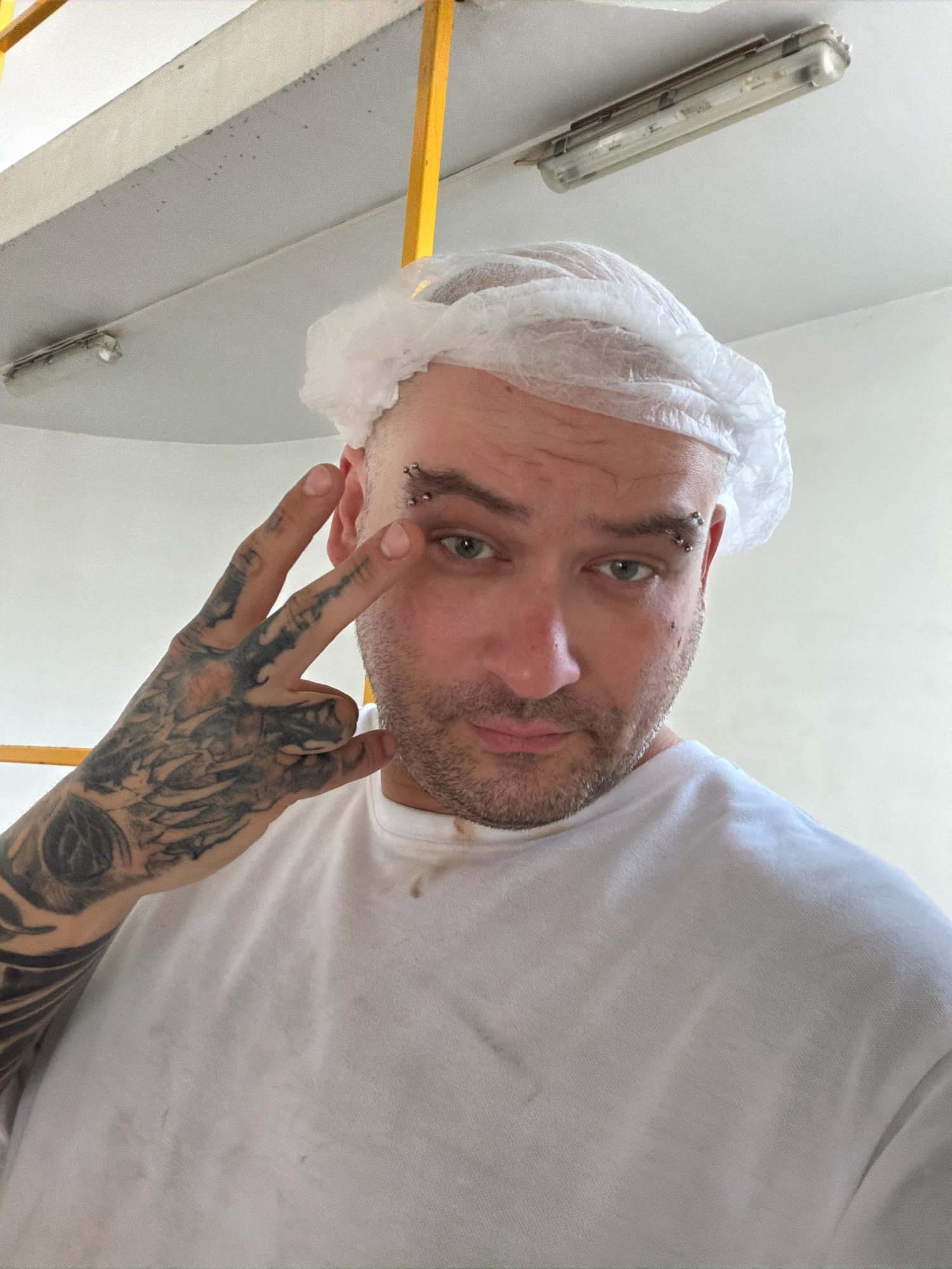A man wearing a white disposable hairnet and a white t-shirt is looking at the camera. He has multiple eyebrow piercings and tattoos on his left arm, which is raised in a peace sign. He appears to be indoors, with a ceiling light and yellow support beams visible in the background. The environment is clean and well-lit, suggesting a workplace or a facility. The man's expression is neutral.