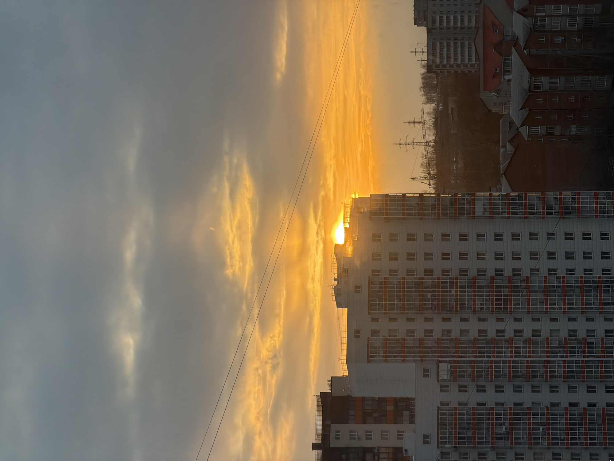 The image shows a cityscape at sunset. A large, modern apartment building with white facade and red window frames is prominent in the foreground. Behind it are other buildings, including a taller apartment block and older, red-roofed structures. In the background, a line of trees is visible, as well as what appear to be the masts of sailboats and electricity pylons. The sun is setting, creating a bright, golden glow that fills the sky. The scene is likely from Krasnoyarsk, Russia, considering the prominent sailboat masts, which are part of a popular attraction on the Yenisei river. Two electrical wires are seen.