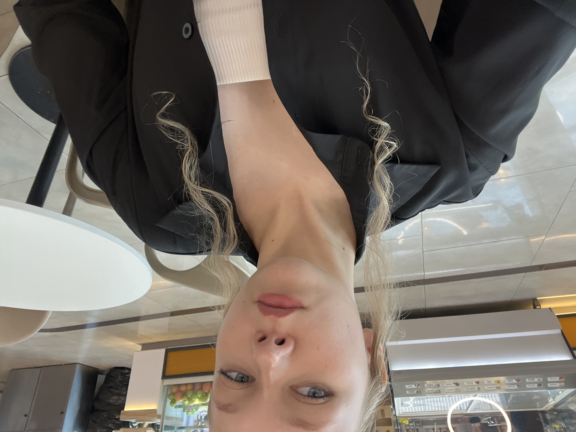The image shows a young Caucasian woman with blonde hair and blue eyes. She is wearing a black collared shirt over a white ribbed top. She is seated at a round white table with a black base, likely in a cafe or restaurant. In the background, there are display cases that appear to contain food items, possibly at a bakery or ice cream shop. The floor is tiled with a reflective surface. The overall setting suggests an indoor commercial establishment. The image does not contain any specific indicators to pinpoint its location within the city of Perm, Russia.