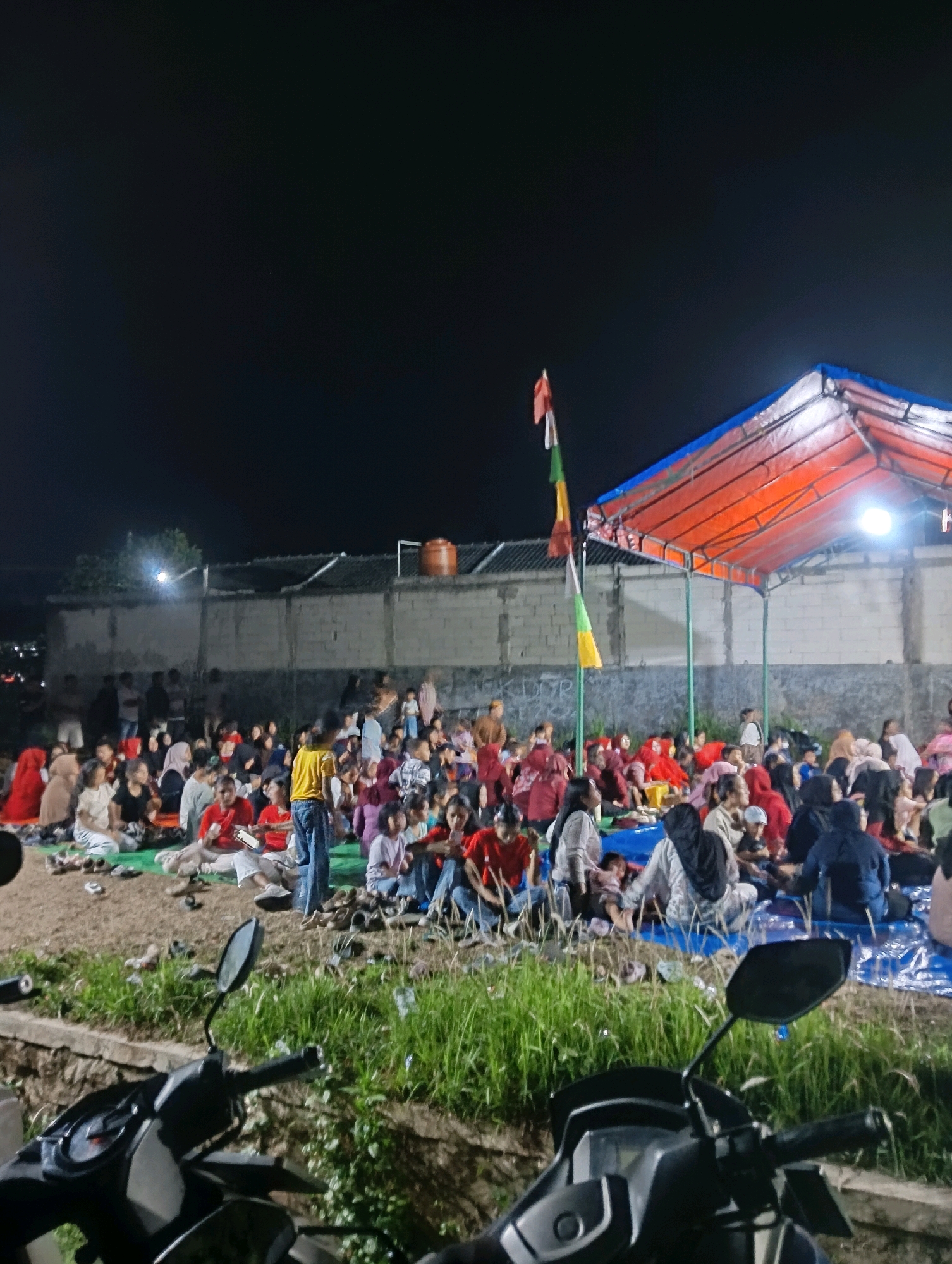 An outdoor nighttime gathering is depicted in Cikasungka, Indonesia. A large group of people, appearing to be a mix of adults and children, are seated on the ground. Some individuals are seated on mats, while others are on blue tarps. A temporary orange tent structure with a blue roof is set up, and flags are visible. In the foreground, parts of two motorcycles are visible, suggesting they are parked nearby. The background shows a low-rise building with a tiled roof and a water tank. The scene appears to be in a somewhat undeveloped area with grass and dirt present. The overall atmosphere suggests a community event or informal gathering.