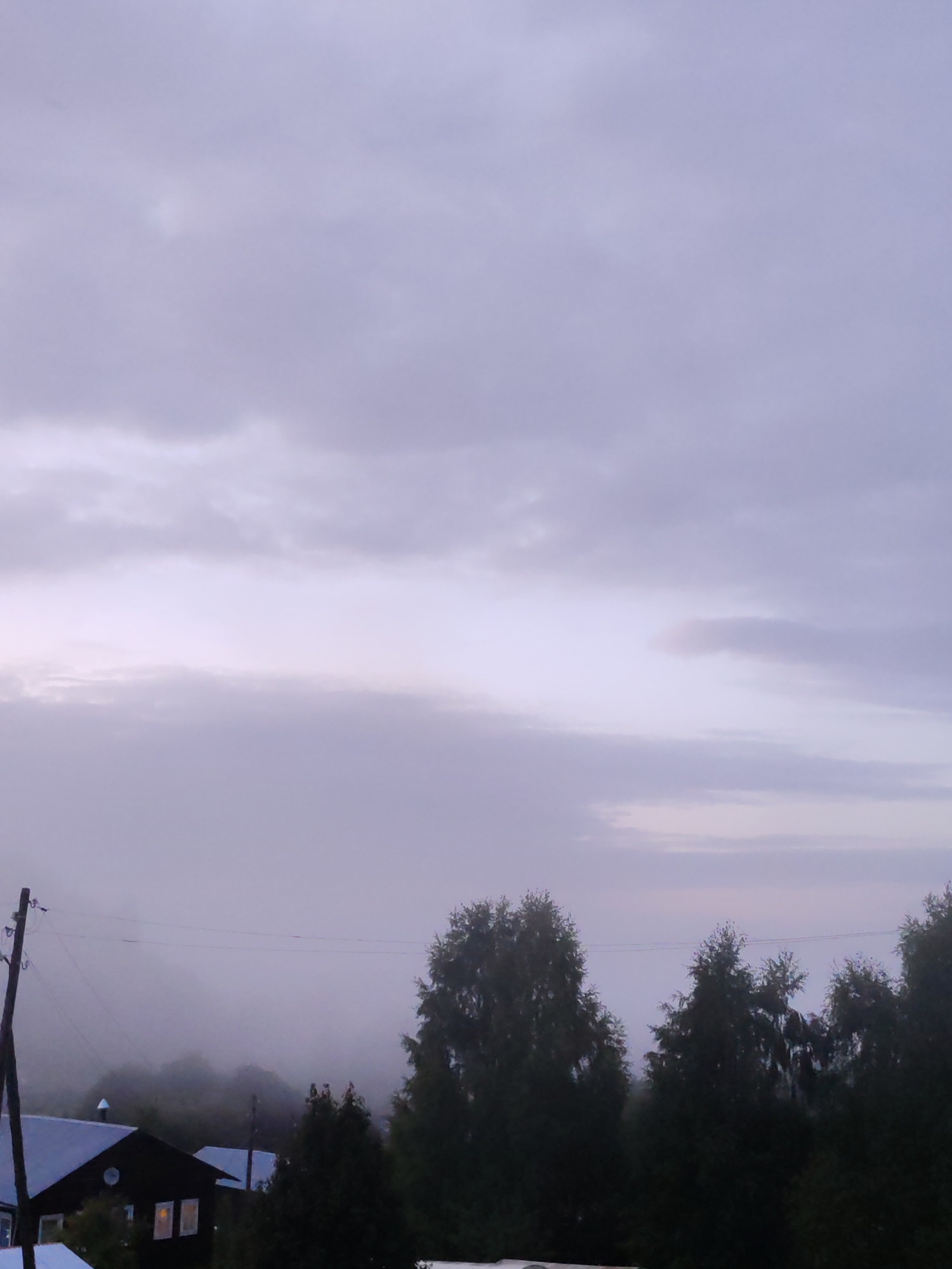 The image depicts a misty, overcast sky above a rural landscape in Чухлэм, Russia. In the foreground, a dark-colored house with illuminated windows is visible. To the left, a utility pole with wires is present. Dense evergreen trees line the middle ground, with a fog-laden background suggesting a natural or hilly terrain. The overall atmosphere is subdued due to the cloudy weather and low light conditions.