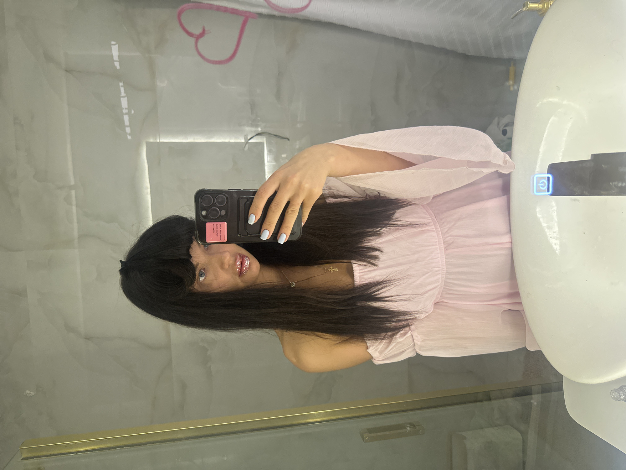 A young woman is captured in a mirror selfie, facing the camera with a neutral to slight smile. She has long, dark hair with bangs, styled straight, and is wearing a light pink, flowing dress or top. Her lips are adorned with glittery gloss, and her fingernails are painted a light blue color. A delicate necklace with a small cross pendant is visible around her neck.
She holds a black smartphone, featuring multiple cameras on the back, in her right hand. A pink sticker is attached to the phone case, on which fragmented text, possibly reading "CIEAR ME MYTO YOUR WORTHINESS," can be discerned due to reflection and angle.
The setting appears to be a modern bathroom, likely in Moscow, Russia, characterized by white marble-patterned walls reflected in the mirror. A portion of a white sink is visible on the right side of the frame, with a small gold-colored fixture or tap above it. Below the tap, a blue illuminated power symbol icon is visible on the sink or mirror surface. A pink heart-shaped object, possibly a decoration or hanger, is reflected in the upper left portion of the mirror. A gold-colored ledge or frame runs along the bottom edge of the mirrored surface. The lighting is bright, suggesting an indoor, well-lit environment.