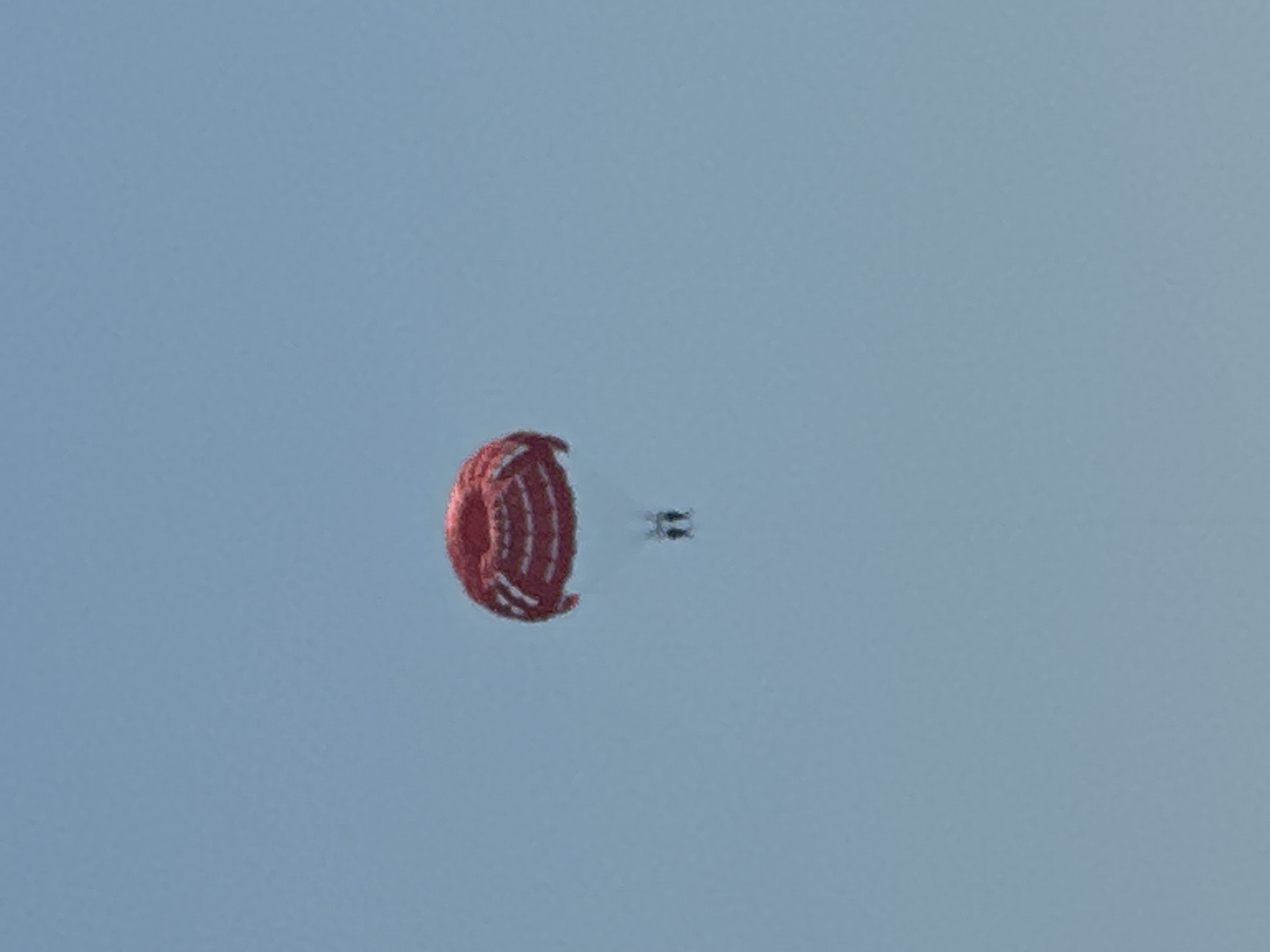 A red and white parachute is suspended in a clear blue sky, with two figures visible beneath it, appearing to be parasailing. The image is shot from a low angle, looking upwards. The overall scene is one of recreational activity in an open, aerial environment.
