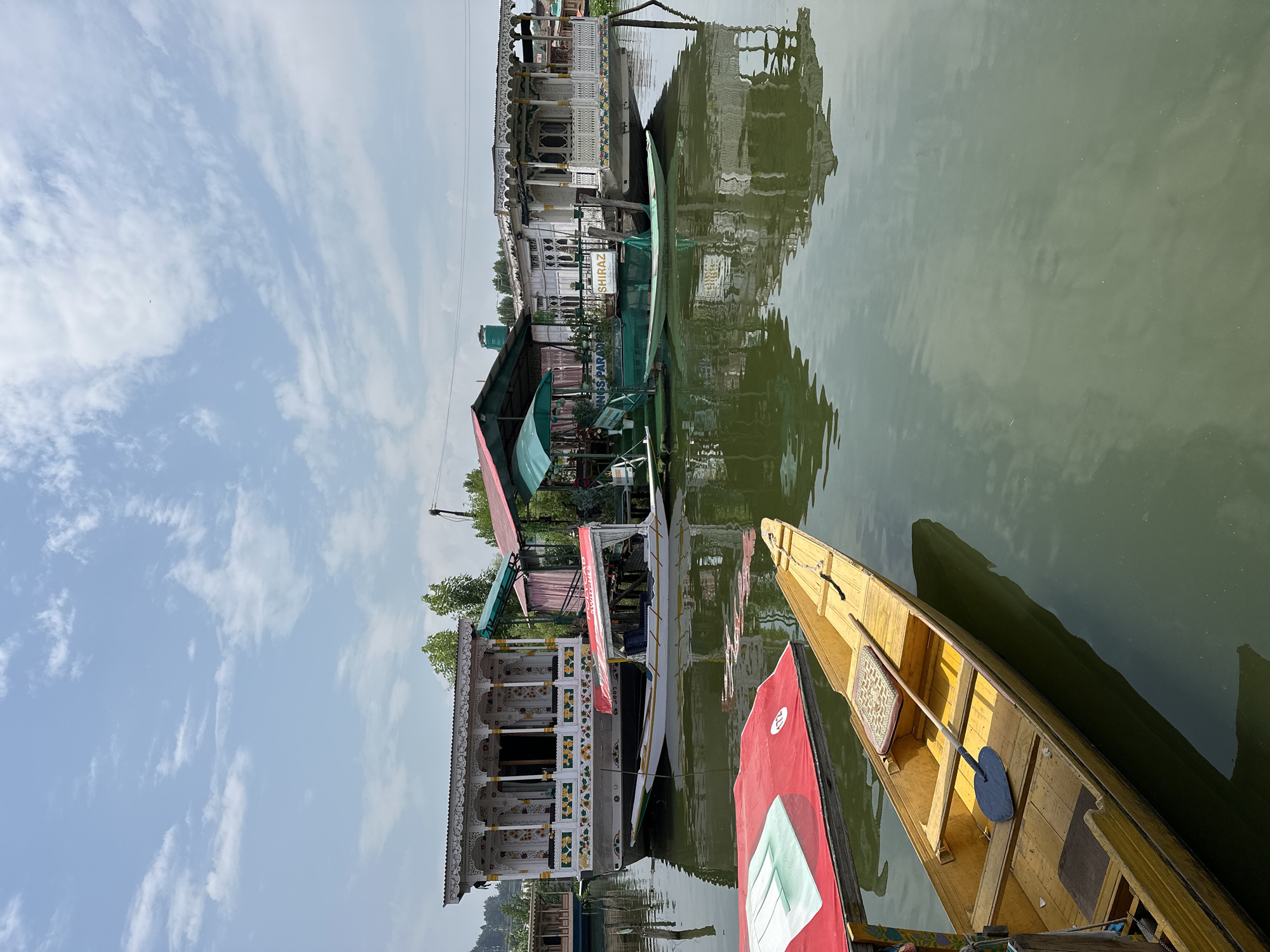 A bright, slightly overcast day illuminates the tranquil waters of Dal Lake in Srinagar, India. Multiple houseboats, their ornate facades reflecting in the water, are moored together. The largest, a white structure with intricate carvings and colorful floral accents, dominates the background. In the foreground, two smaller boats are visible. The closest is a bright yellow, traditional Shikara, with a padded seat and a large paddle resting within. Partially obscured by this Shikara is another boat with a red canopy. The water is a muted green, disturbed only by the gentle ripples around the boats, creating soft reflections of the houseboats and the sky. Green trees dot the horizon behind the houseboats.