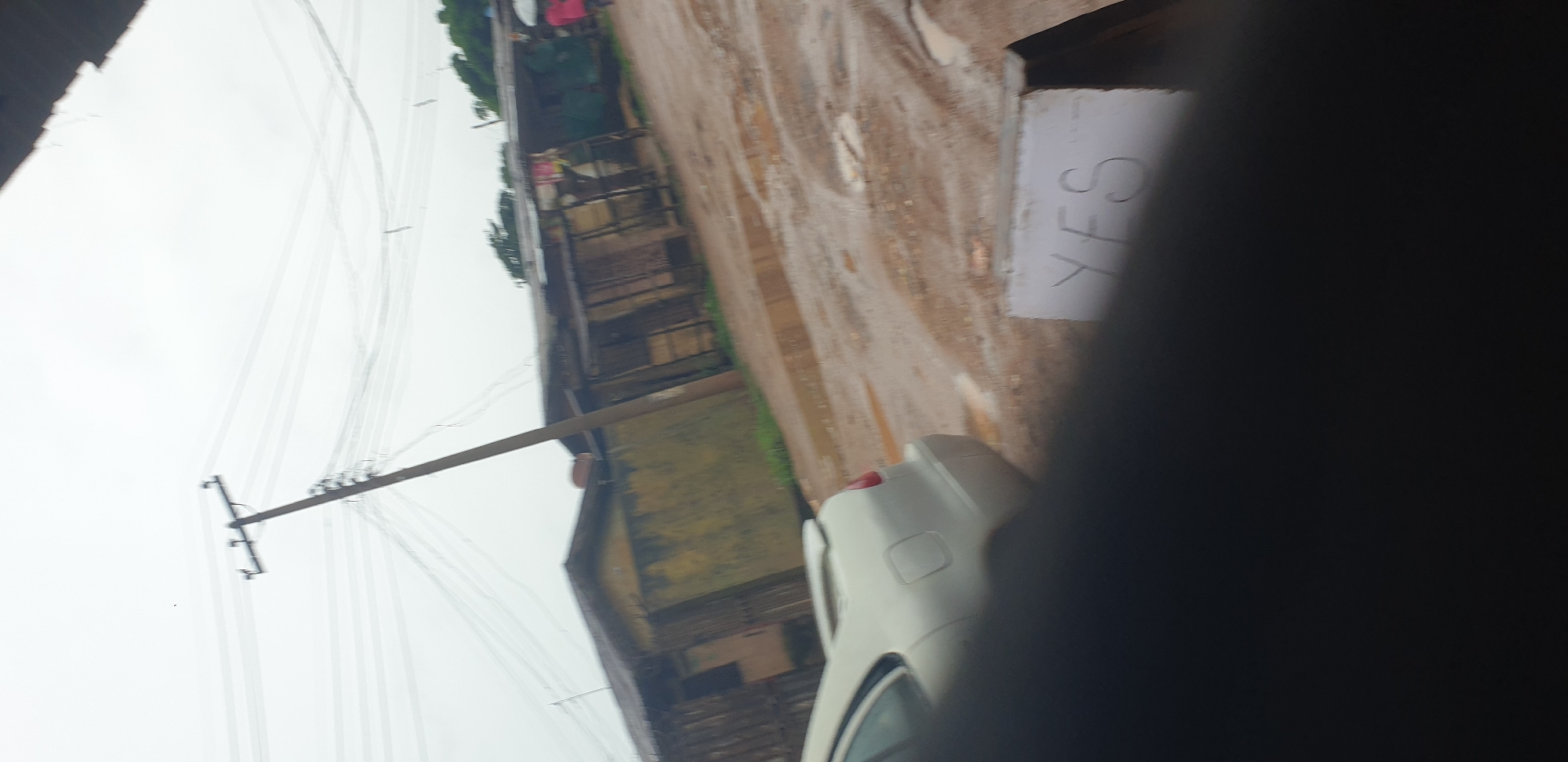 A tilted, low-angle shot captures a scene in Sabon Tasha, Nigeria, under an overcast sky. In the foreground, a portion of a light-colored car is visible, with its rear bumper and spoiler facing left. To the right, a white sign with the word "YES" scrawled in dark lettering is placed on the ground, partially obscured by the car and darkness. The ground is muddy and wet, with several puddles reflecting the dull light. In the midground, a row of dilapidated buildings with corrugated metal roofs and wooden walls stretches across the frame, appearing weathered and aged. A utility pole with multiple wires extends upwards on the left side of the image, disappearing into the hazy sky. In the distance, more buildings and some greenery are faintly visible. The overall impression is one of a rural or semi-urban environment, possibly affected by recent rainfall.