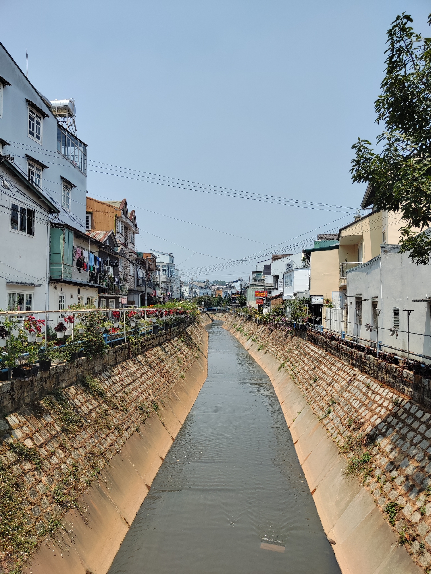 The image shows a canal flowing through the city of Cam Ly - Đà Lạt, Vietnam. The canal has concrete-lined sides and contains murky water. The banks of the canal are lined with stone blocks, and there are plants in pots along the sides of the canal. Buildings are situated on either side of the canal, including houses and shops. Some of the houses have clotheslines with laundry hanging out to dry. Power lines run above the canal. The sky is clear and blue.