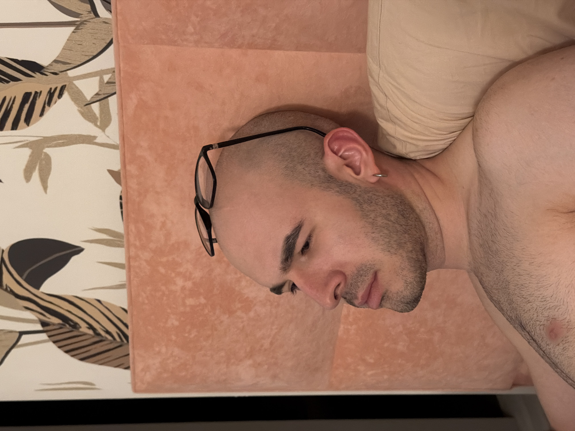A close-up, head-and-shoulders shot depicts a bald man with a trimmed beard and glasses resting on his head. He is lying on a peach-colored cushioned surface and a neutral-toned pillow. A leaf-patterned wallpaper is visible on the left. There is no information to identify the location as Kazan, Russia.