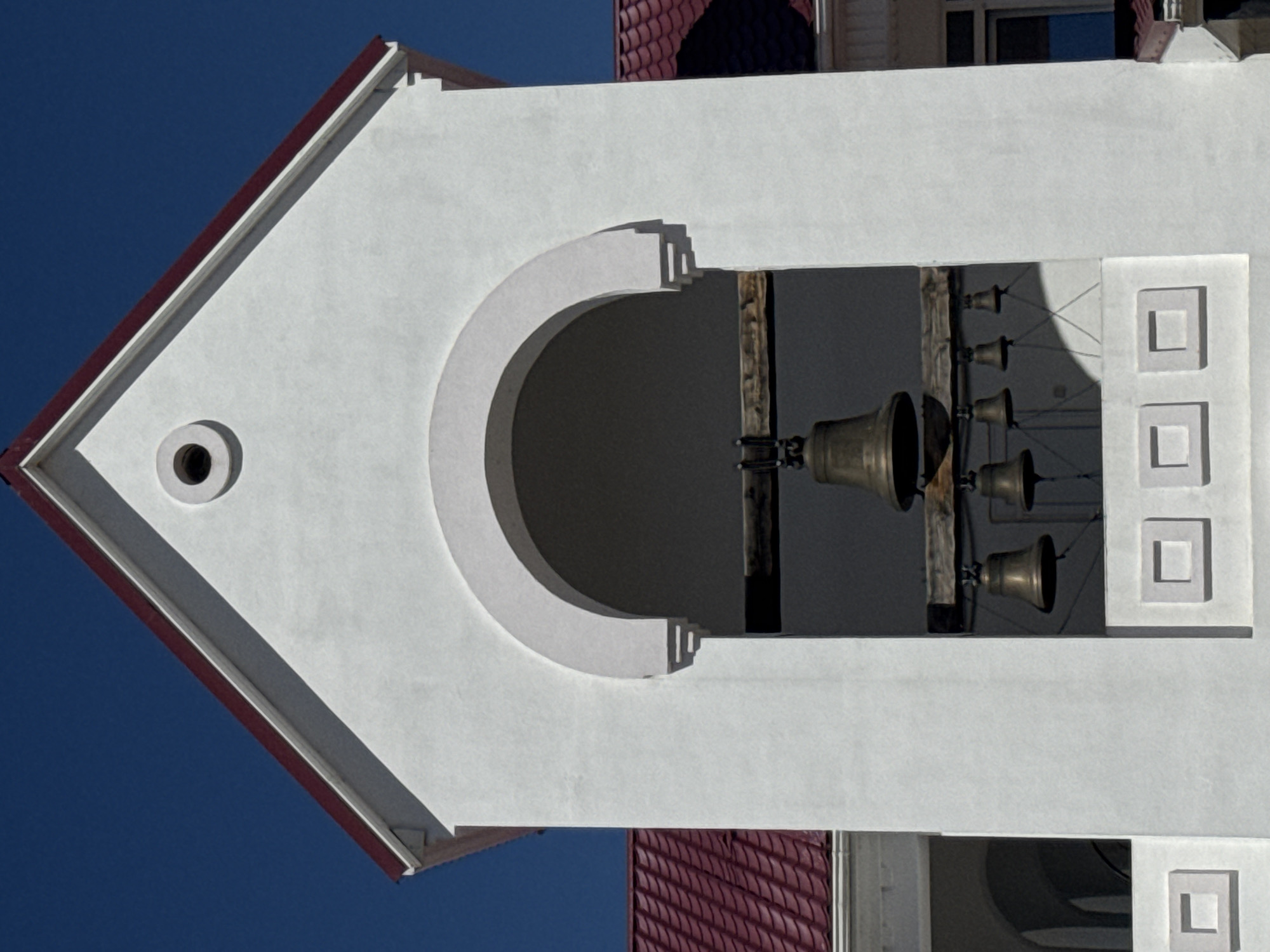A view of a church steeple in an unknown city shows a white facade with a red trim and a hole, a large arched window, and a window with several bells inside.