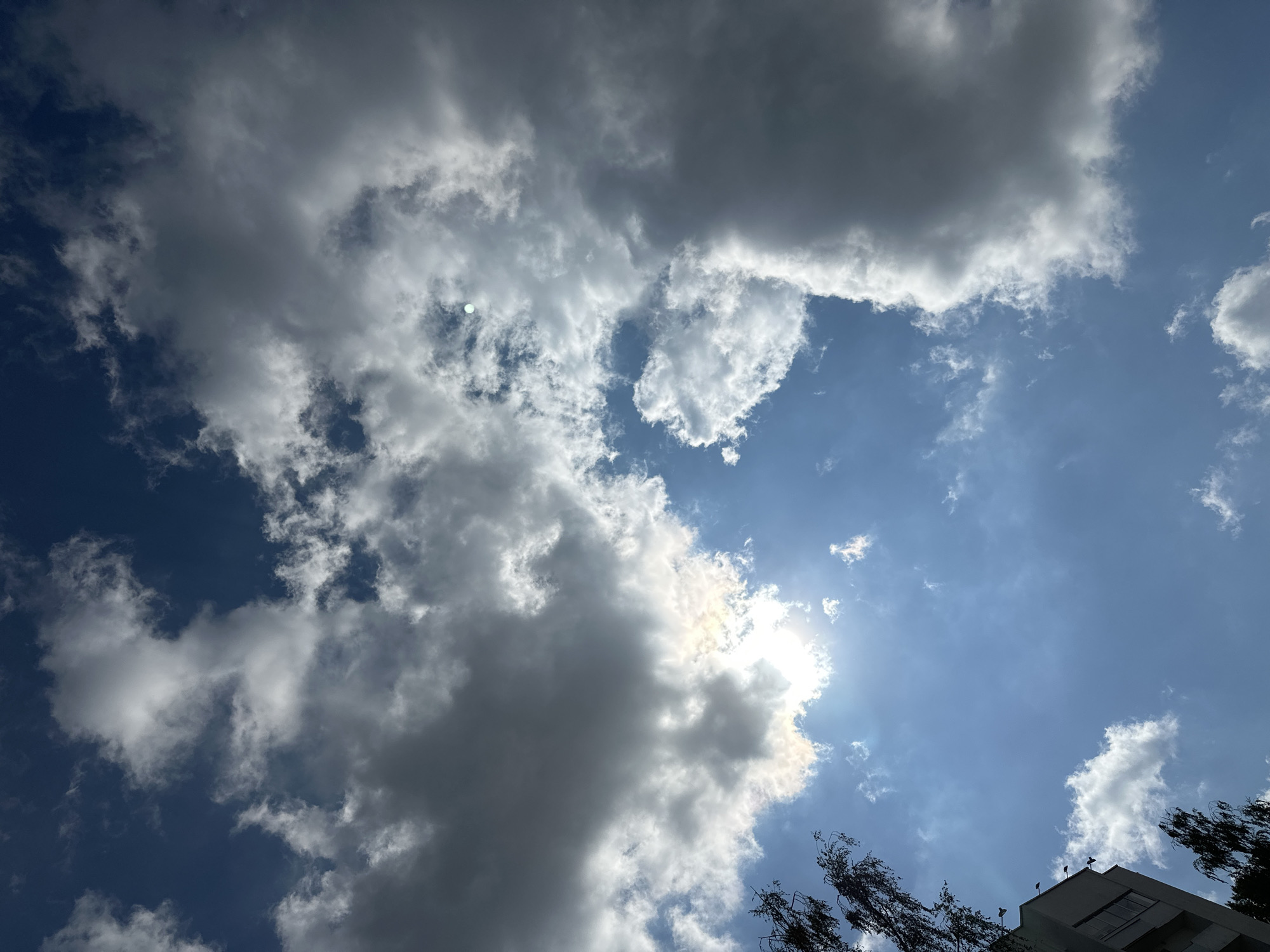 The image captures a sky filled with a mix of dark and white clouds. The sun is visible, peeking through the clouds and casting a bright light. The lower left portion of the image shows a building with windows, and trees with dark branches are visible on the left and lower edges. The sky is a deep blue. The scene is likely an outdoor view from a city, possibly Minsk, Belarus, given the context.
