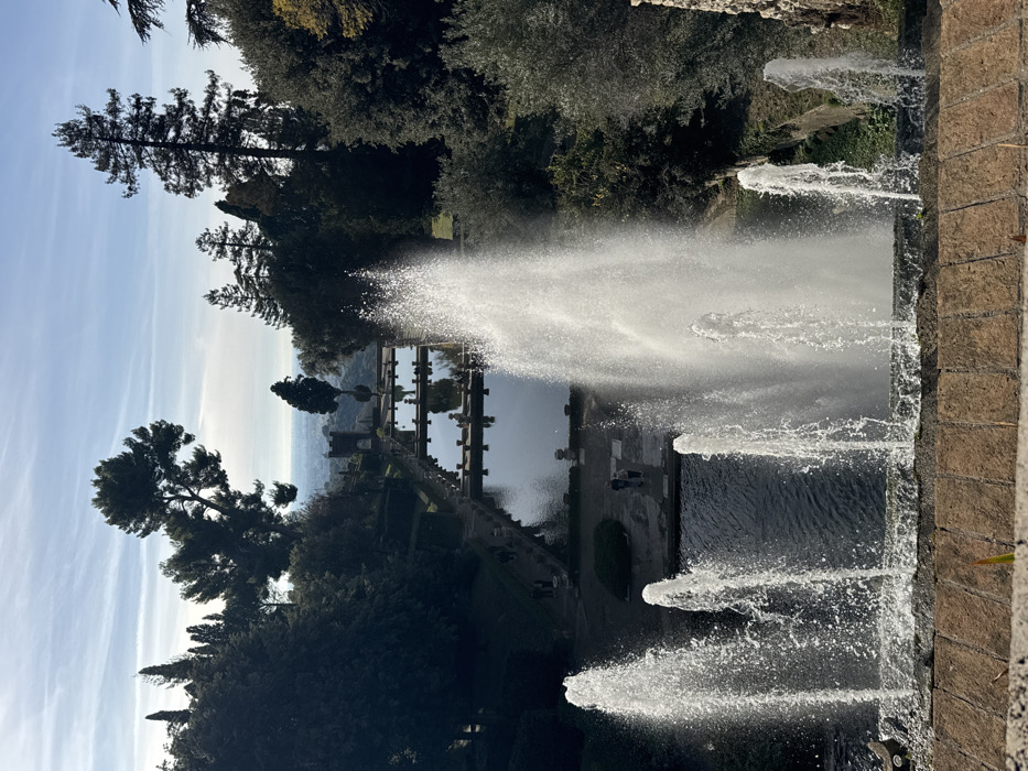 This image captures a descending view within the terraced gardens of Villa d'Este in Tivoli, Italy. In the immediate foreground, five powerful water jets erupt from a stone basin, with the central spray being the tallest and most voluminous, flanked by two shorter jets on each side. The water is white and appears turbulent due to the force of the spray, catching direct sunlight. Below this, a series of rectangular, dark water pools are arranged in a cascading terraced formation. Stone pathways border these pools, along which approximately ten indistinct human figures are distributed, appearing to walk or stand. Decorative stone urns are positioned alongside the pool edges. The midground and background consist of dense, dark green foliage, including tall evergreen trees such as pines and cypresses, framing the descending landscape. In the far distance, a light blue sky with scattered clouds hovers above a hazy cityscape, and a dark stone tower with arched openings is visible within the lower garden sections. The scene is illuminated by clear daylight.