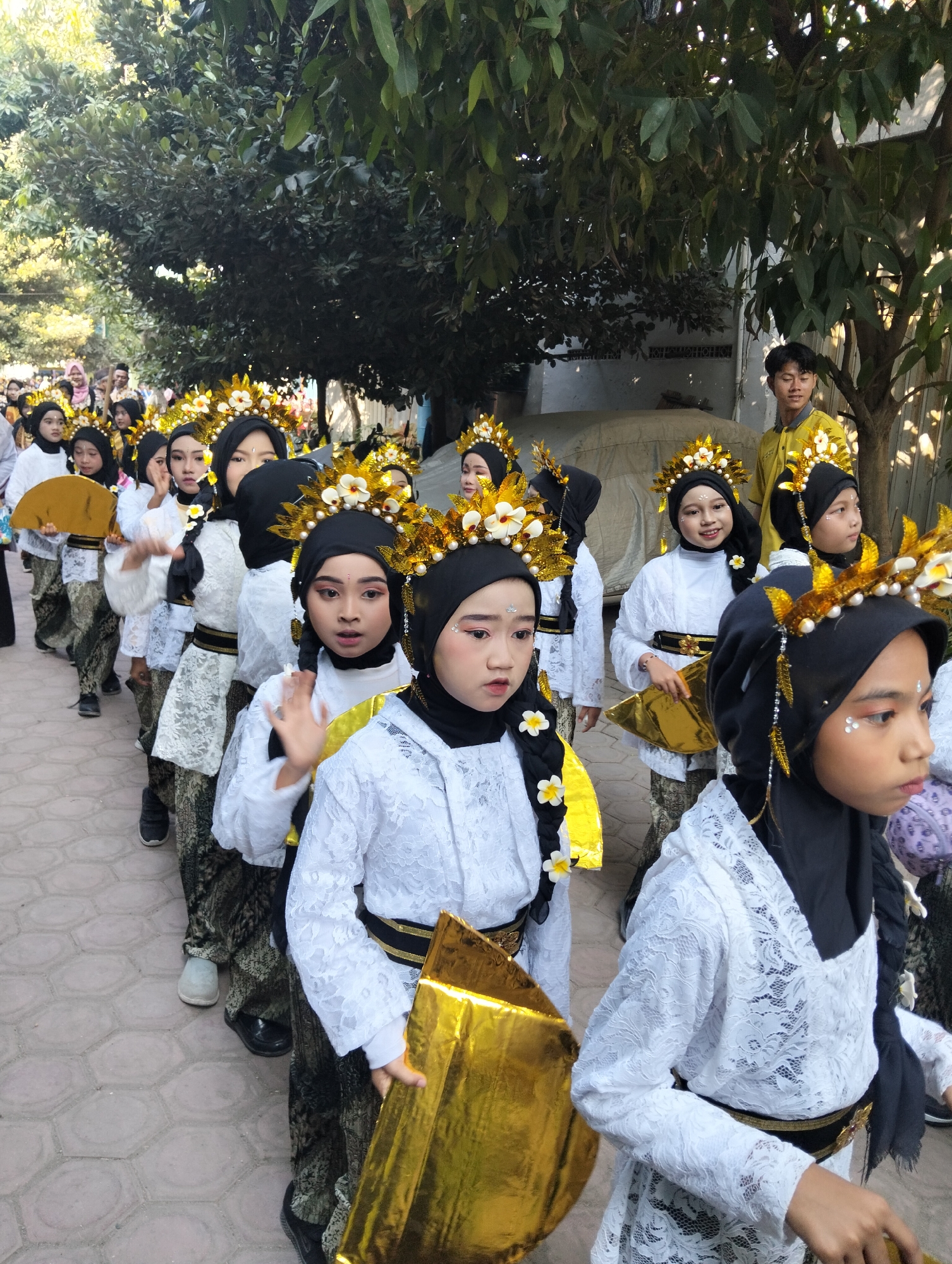 A group of young girls in traditional Indonesian attire are marching in a procession in Kranji, Indonesia. The girls are wearing white tops with gold headpieces and black hijabs, and patterned pants. They are holding gold props. The procession is taking place outdoors on a paved path lined with trees. In the background, a young man in a yellow shirt is visible. The lighting suggests it is daytime.