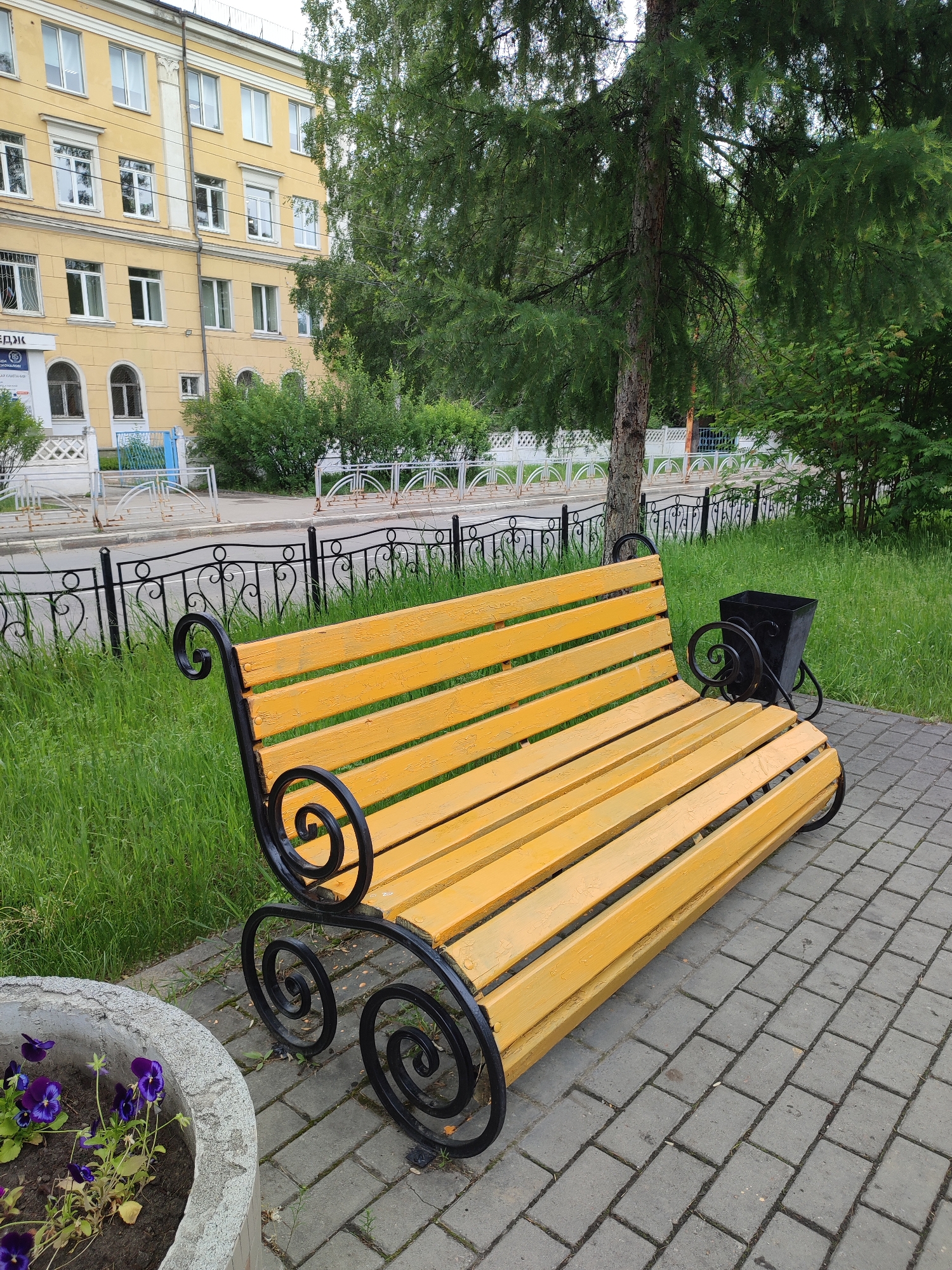 A yellow wooden bench with ornate black metal scrollwork supports sits on a gray brick pathway in front of a grassy area and a black metal fence. To the left of the bench, a gray concrete planter holds several purple flowers. In the background, a multi-story yellow building with numerous windows is visible, along with trees and bushes. A black metal trash receptacle stands behind the right side of the bench. The scene appears to be a public space within the city of Angarsk, Russia, likely a park or a pedestrian area adjacent to a building. No people are present.