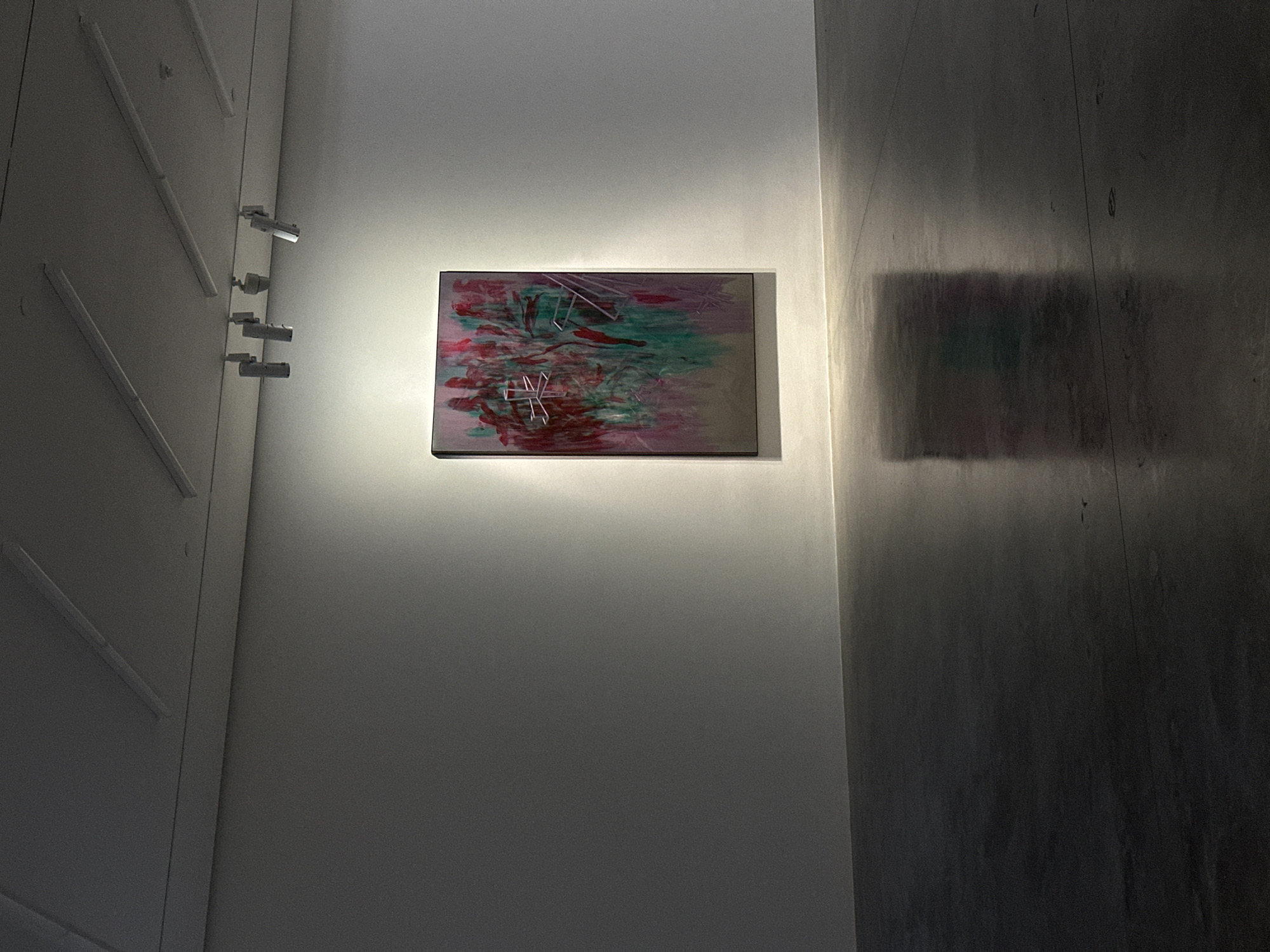 An indoor art gallery space in the city of London, United Kingdom, displays a single, vertically oriented abstract painting mounted on a plain white wall. The artwork, framed in a thin dark border, features vertical streaks of deep red, teal green, and light purple, overlaid with angular white or light-toned linear elements. The painting is brightly illuminated by multiple track-mounted spotlights on the ceiling, casting a distinct rectangular glow around it on the wall. The ceiling also contains parallel rows of linear fluorescent light fixtures. The floor is dark, flat, and highly reflective, mirroring a blurred image of the illuminated artwork and the bright wall area below. No individuals are present in the scene.