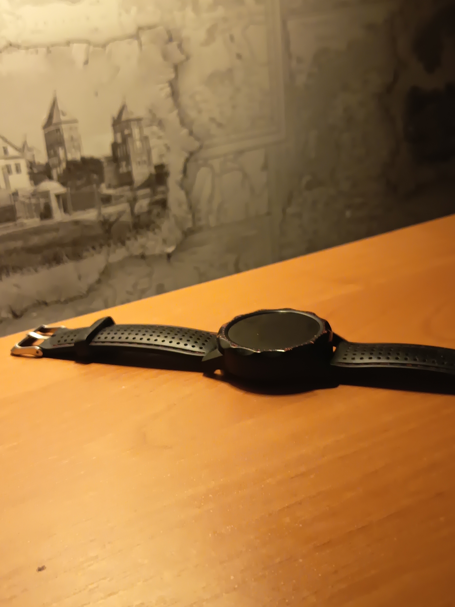 The image shows a black digital watch lying on a wooden table. The watch has a round black face and a perforated black band with a buckle. The background of the image features a wall with a faded mural of buildings. The scene is lit by a warm light, creating shadows on the table and the wall. The location is the city of Izmail, Ukraine.